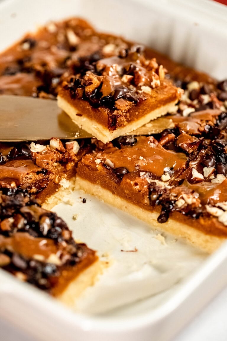 A baking dish with sliced dessert bars topped with caramel, chocolate, and nuts. One piece is being lifted by a spatula.