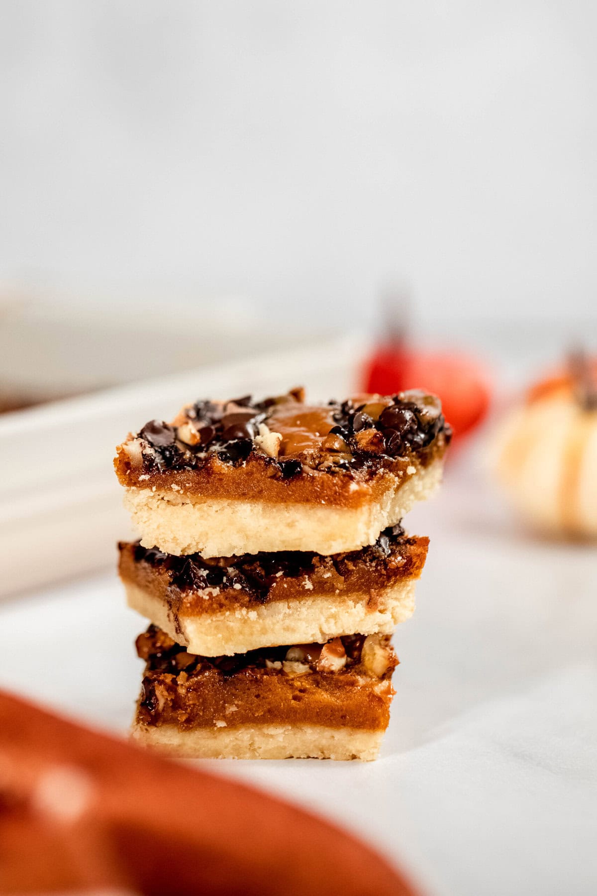 A stack of three dessert bars with chocolate chips and nuts on a light surface. In the background, there is a blurred pumpkin decoration.