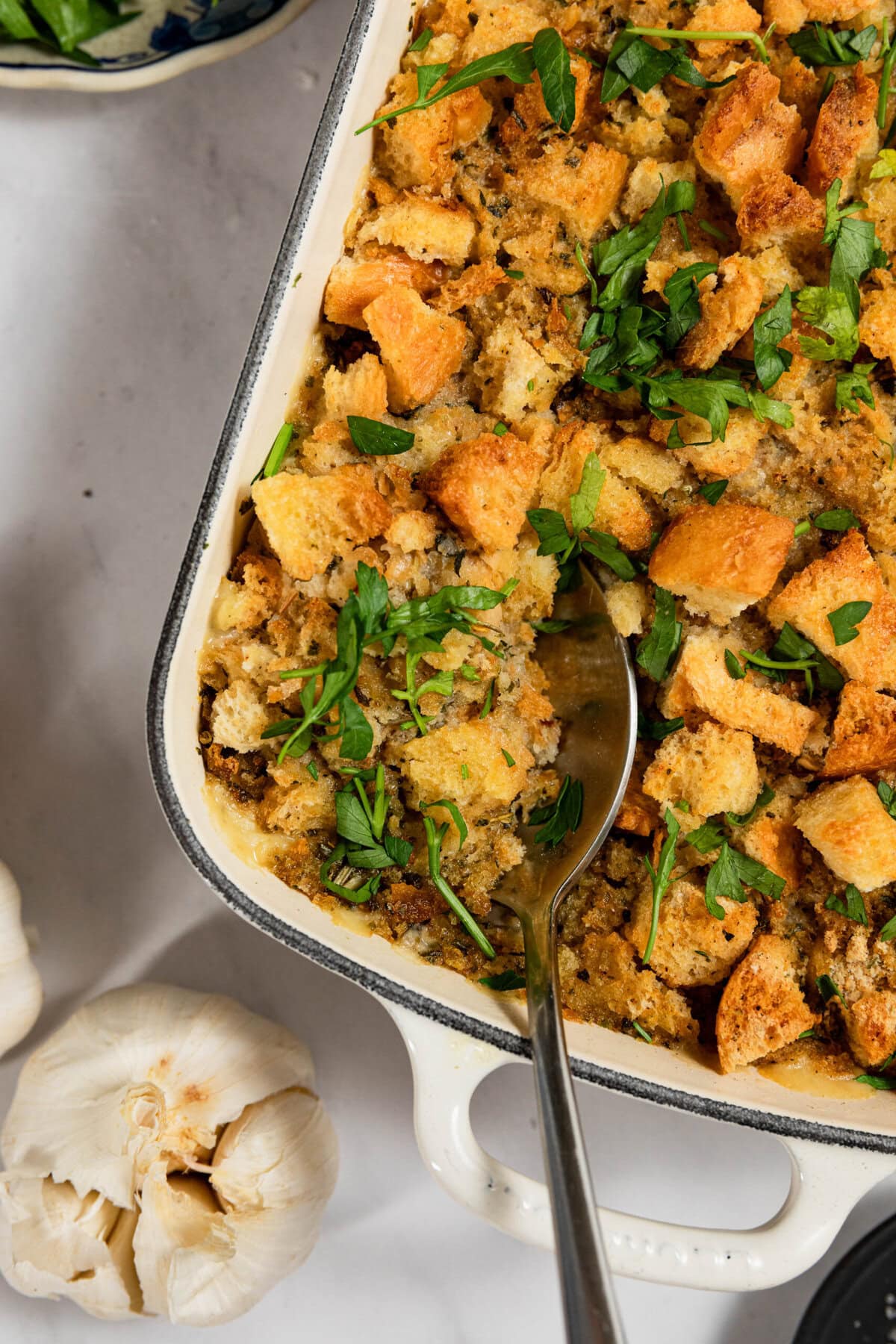 A casserole dish with baked stuffing topped with fresh parsley. A spoon is placed in the dish. Garlic bulbs are nearby on the marble surface.