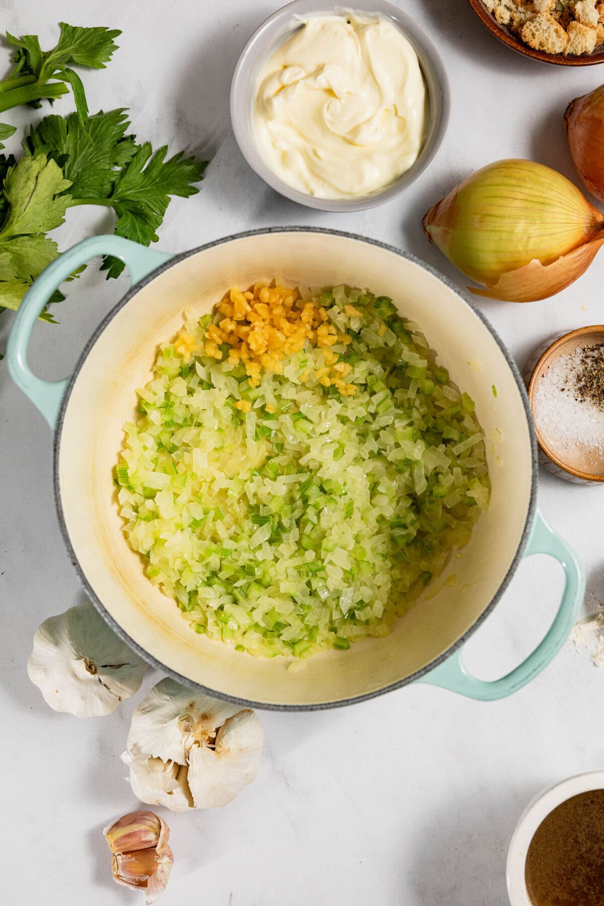 A pot on a white surface containing diced celery, onions, and garlic. Surrounding are ingredients like mayonnaise, breadcrumbs, whole garlic, onion, and spices.