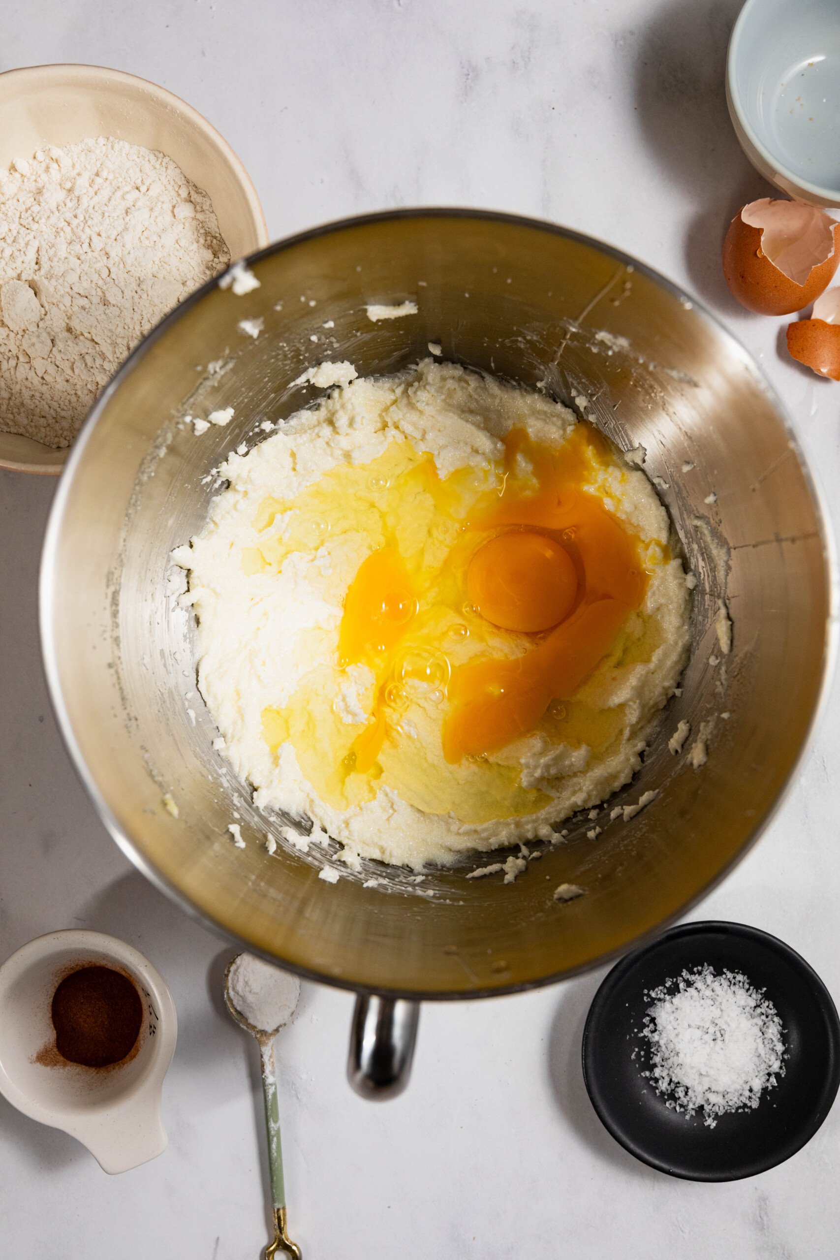 Bowl with cracked eggs on creamed butter and sugar, surrounded by flour, egg shells, cinnamon, and salt on a marble surface.