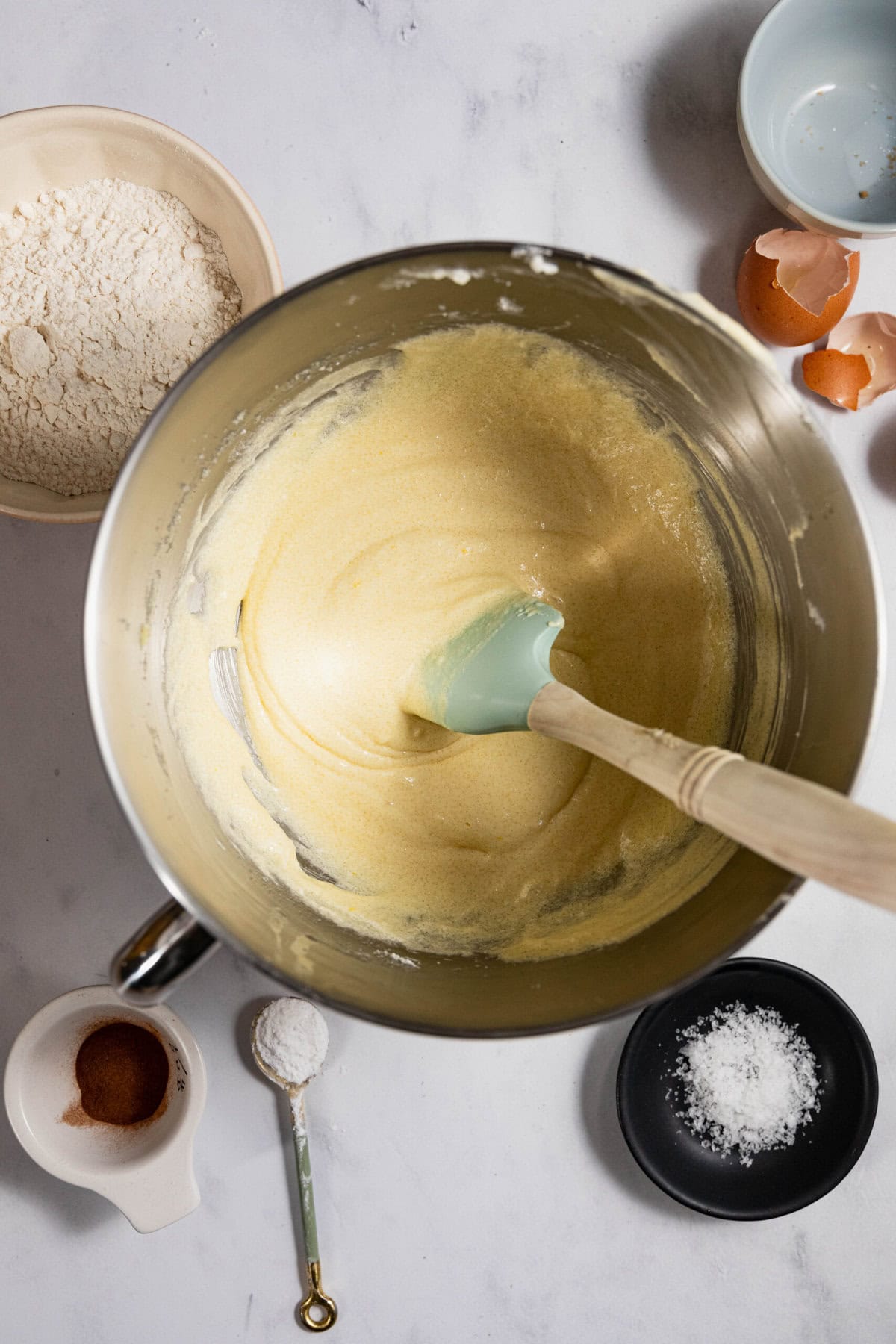 Overhead view of a mixing bowl with batter and a spatula. Surrounding items include flour, eggshells, a blue bowl, a white bowl with cinnamon, a tablespoon, and a black plate with salt.