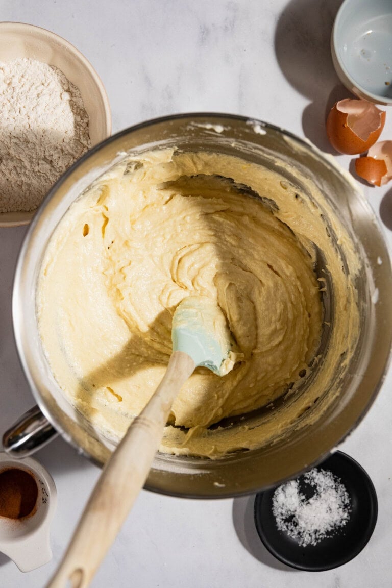 A mixing bowl with yellow batter and a spatula. Surrounding the bowl are ingredients including flour, eggshells, cinnamon, and salt.