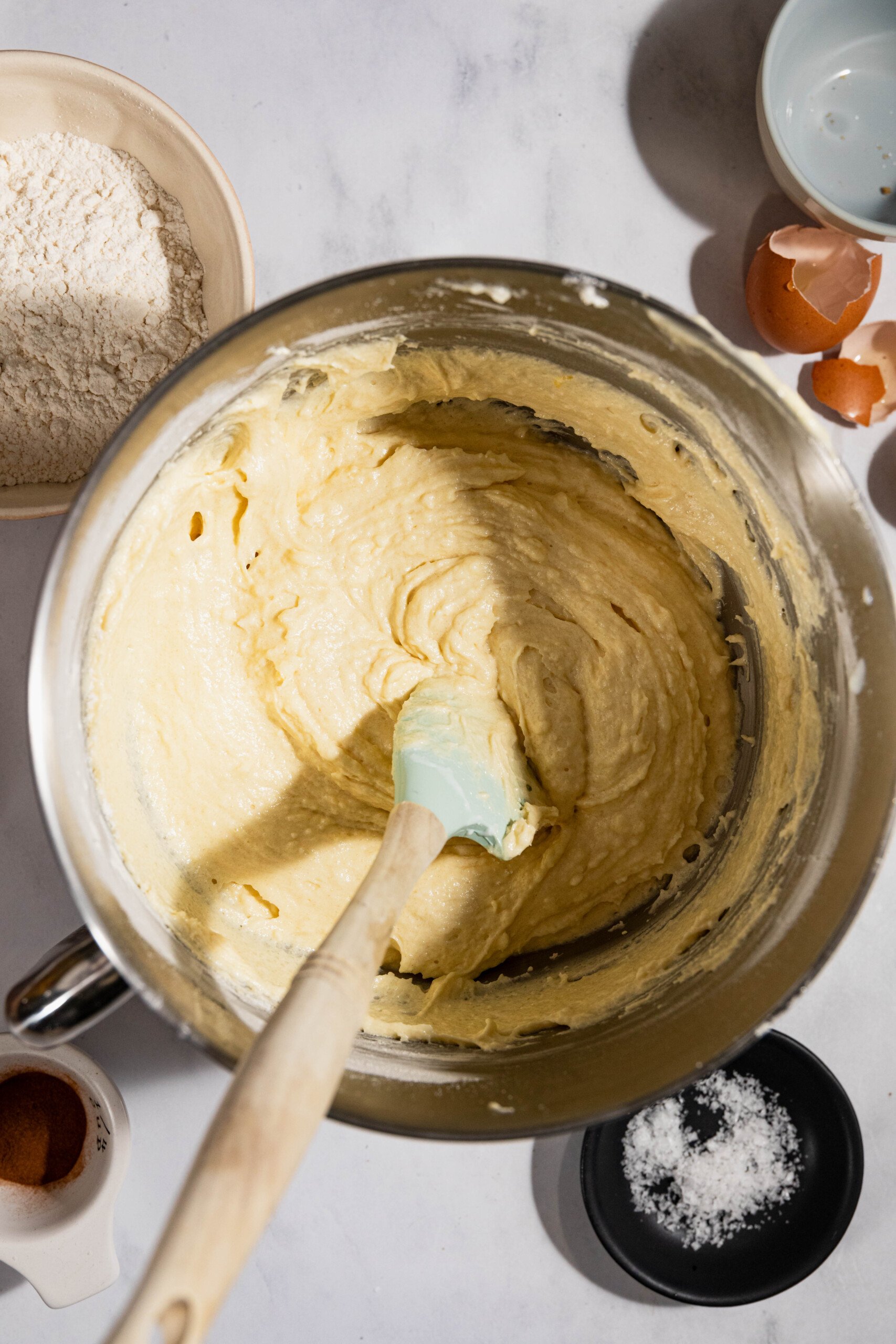 A mixing bowl with yellow batter and a spatula. Surrounding the bowl are ingredients including flour, eggshells, cinnamon, and salt.