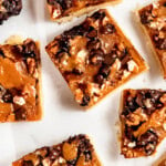 Close-up of dessert bars topped with chocolate chips, nuts, and caramel on a white background.