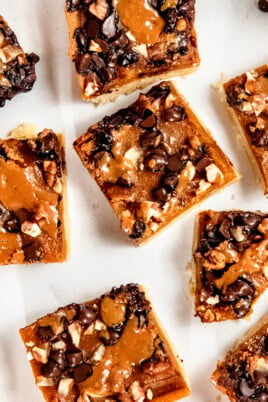 Close-up of dessert bars topped with chocolate chips, nuts, and caramel on a white background.