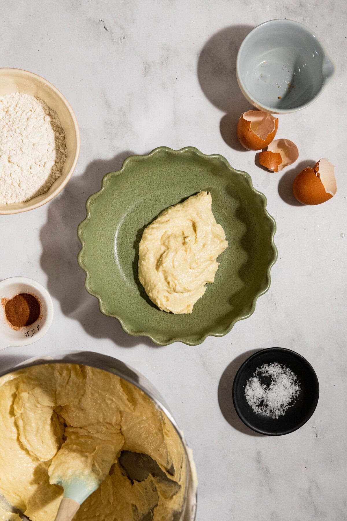 Baking ingredients on a marble surface: flour, cinnamon, cracked eggshells, and salt surround a mixing bowl and a green dish with dough.