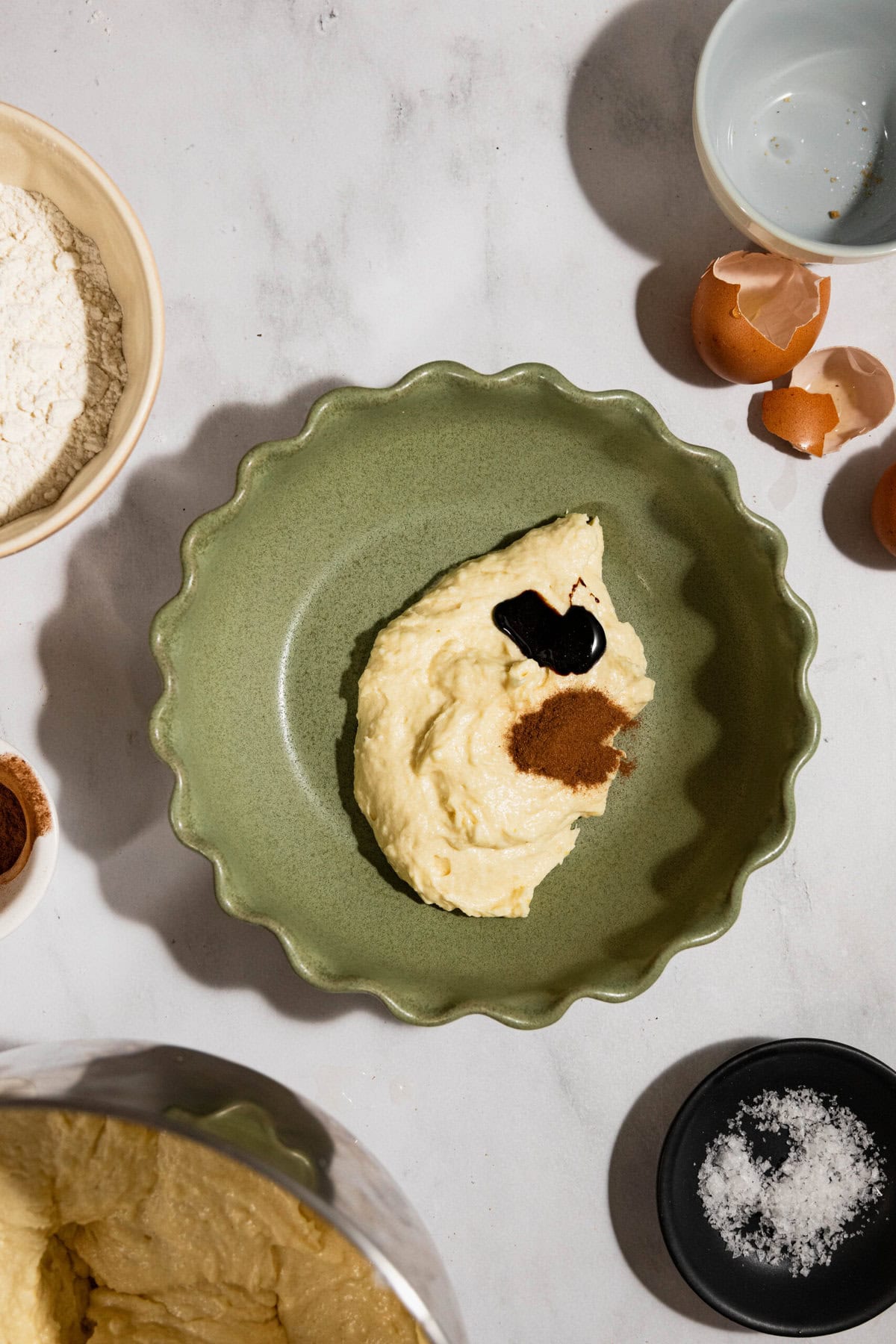 Bowl with batter, vanilla extract, and cinnamon on a counter, surrounded by flour, eggshells, and salt.