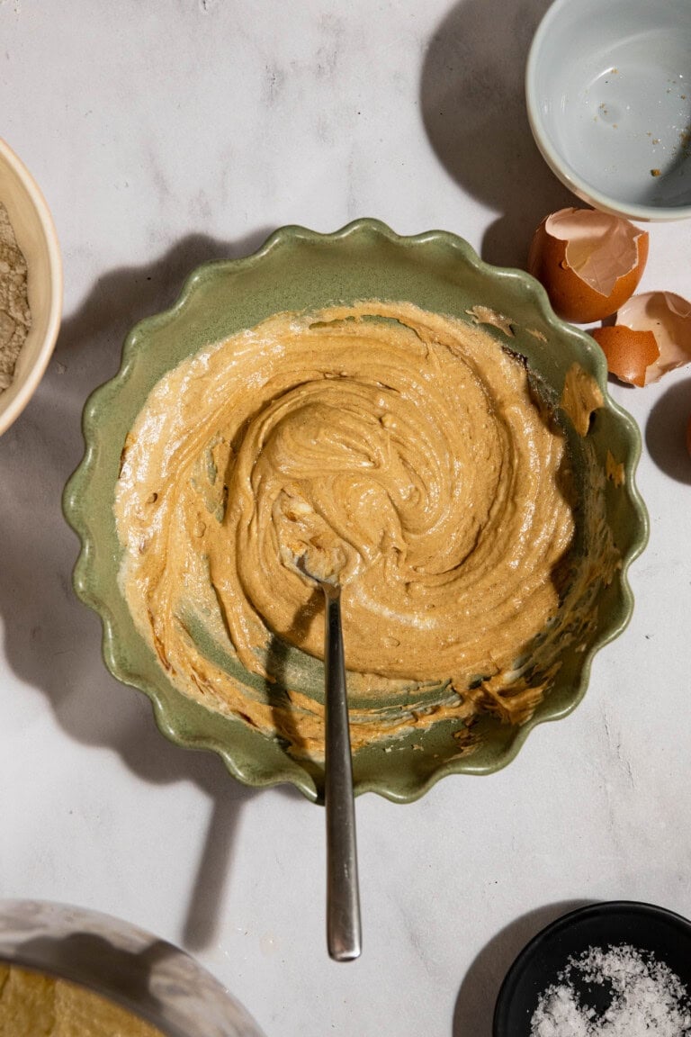 A green bowl with a creamy brown mixture and a spoon. Eggshells, a blue bowl, and a black dish with salt are nearby.