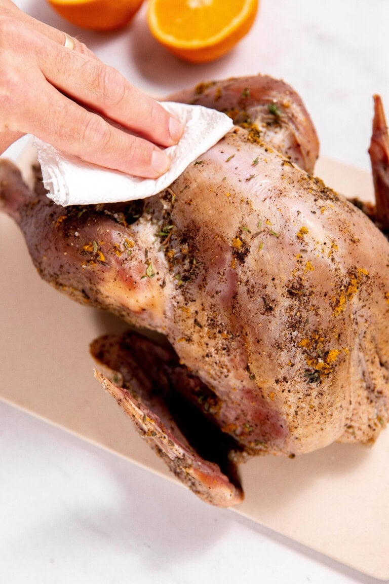 A person is expertly dry brining a raw turkey with an array of herbs and spices on a cutting board. A sliced orange is visible in the background, hinting at the burst of flavor to come.