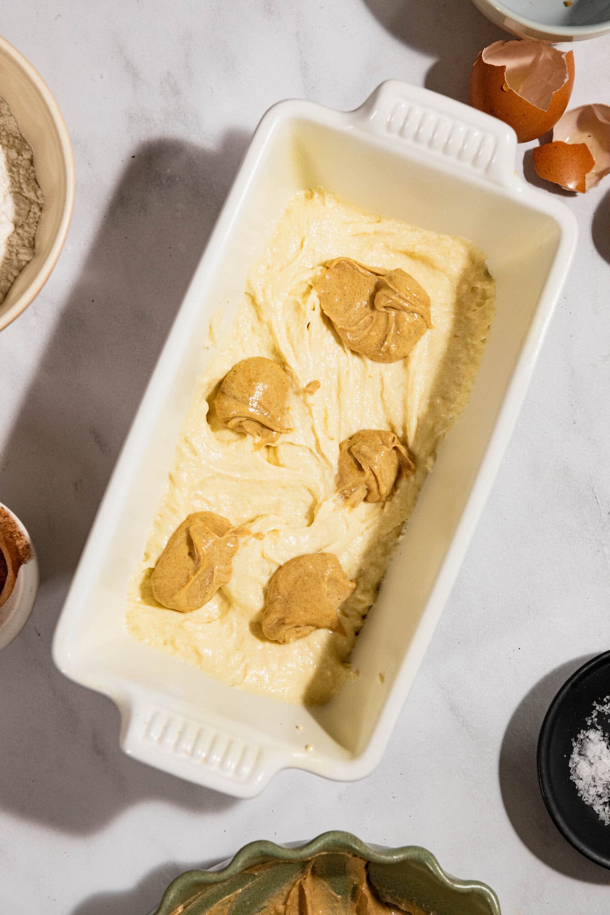 Baking dish with cream-colored batter topped with blobs of a lighter brown mixture. Surrounded by eggshells and bowls of ingredients on a marble surface.