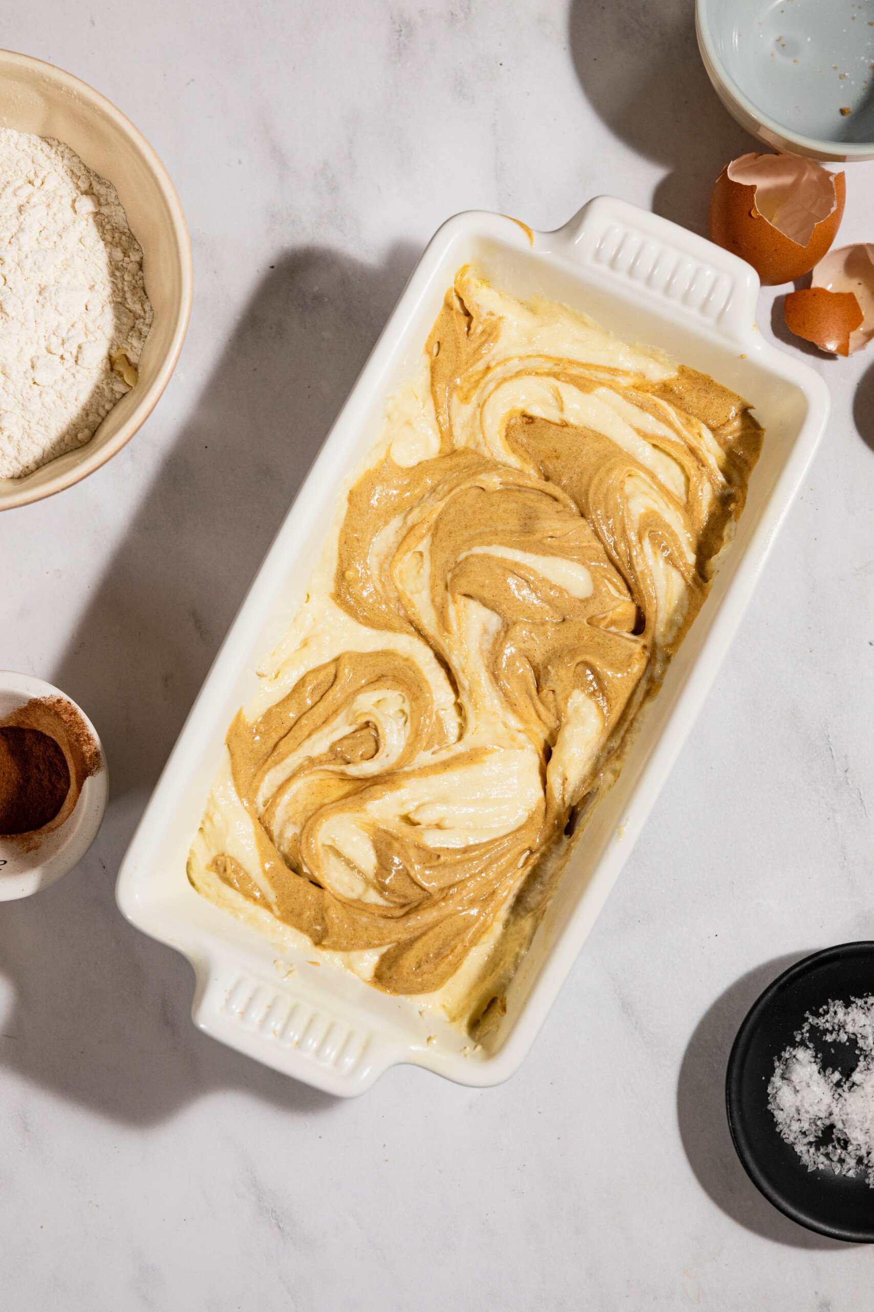 A marble cake batter in a rectangular white baking dish surrounded by bowls of flour, cinnamon, and broken eggshells on a marble surface.