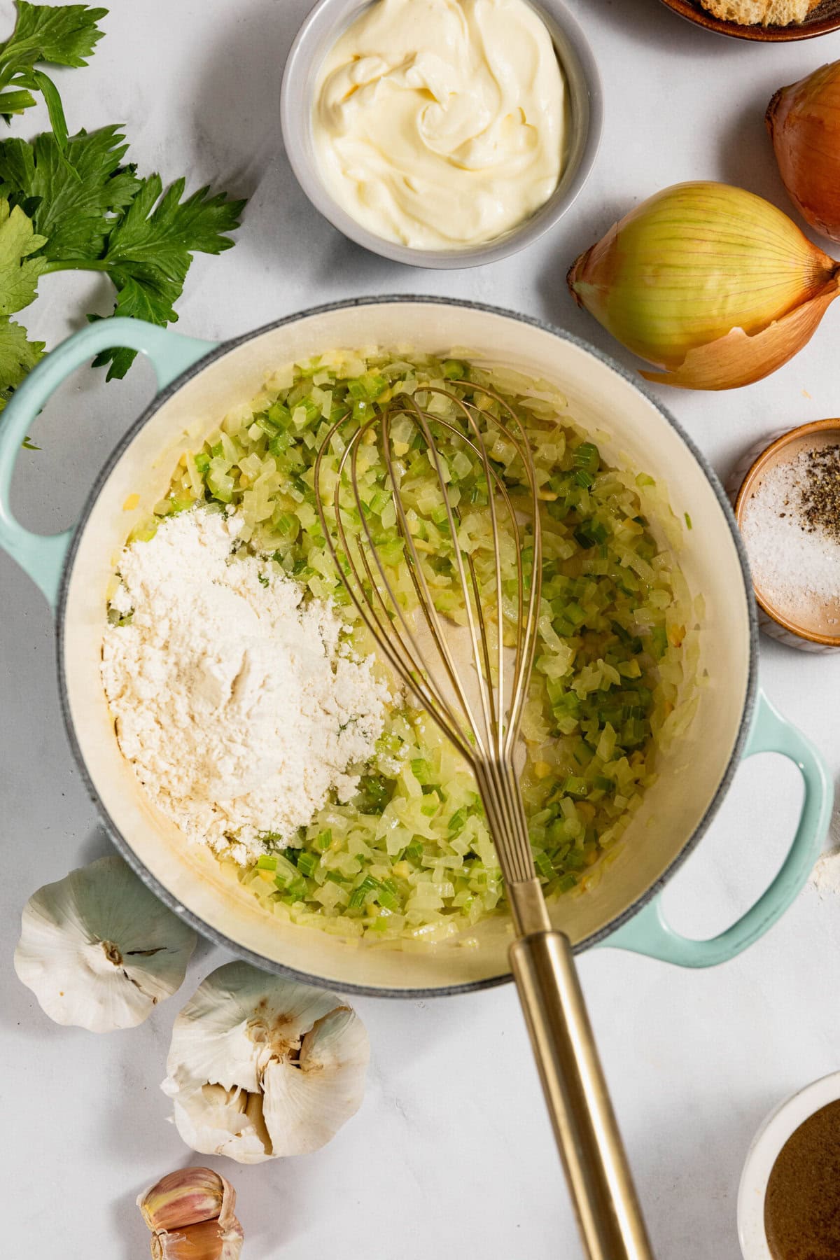 A pot with sautéed onion and celery, a whisk, flour, and nearby ingredients like garlic, onion, parsley, and butter on a white surface.