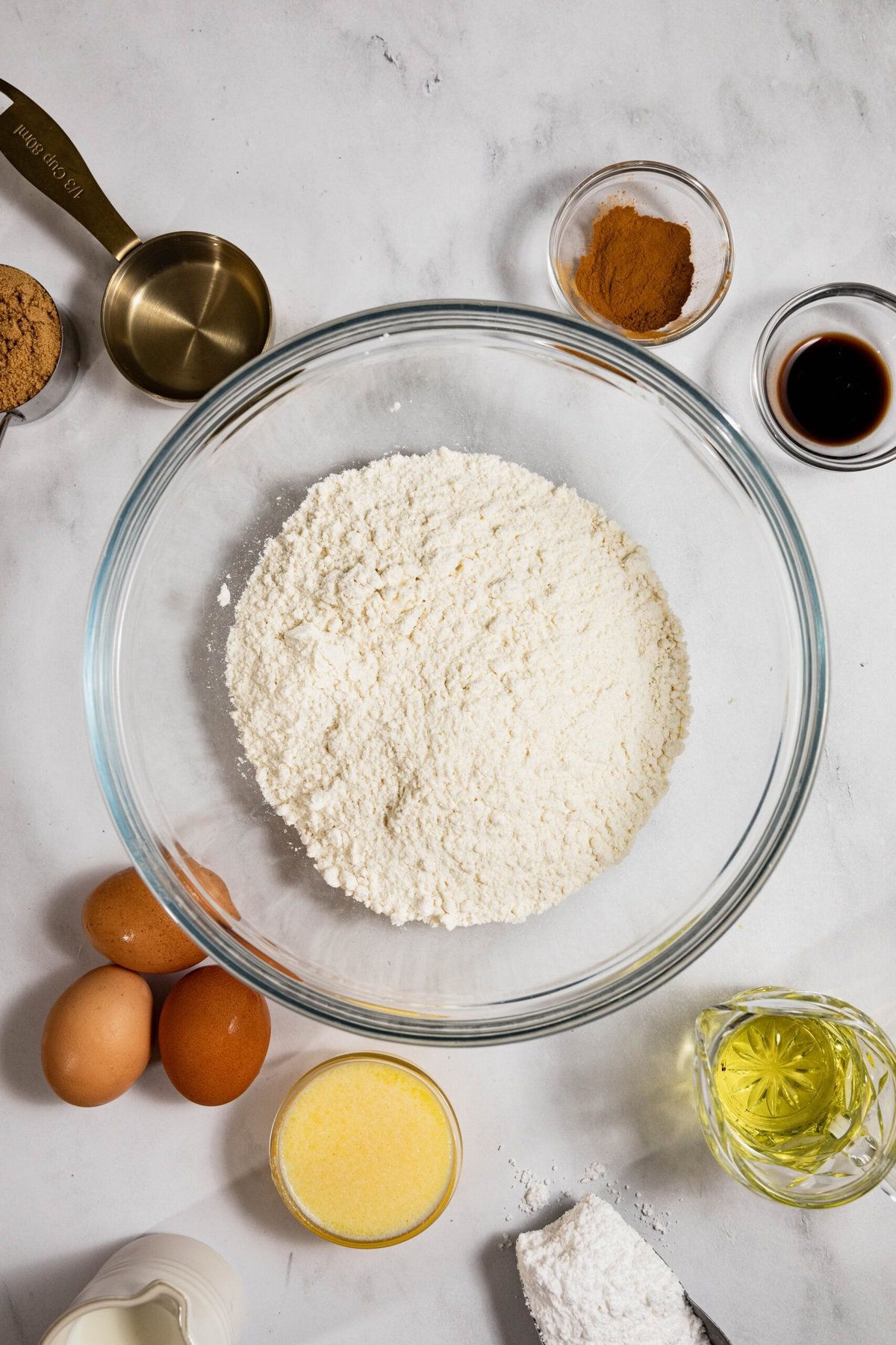 Baking ingredients on a countertop include flour in a bowl, eggs, melted butter, oil, vanilla, cinnamon, and brown sugar. Measuring cup and a small bowl of powdered sugar are also visible.