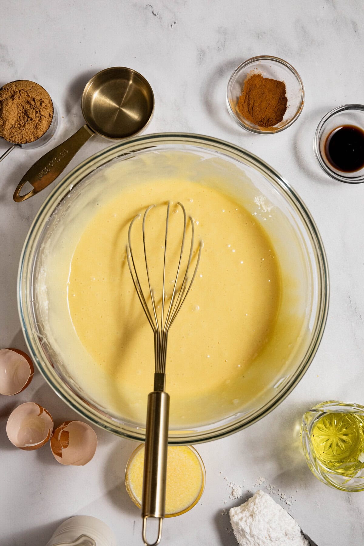 A bowl of yellow batter with a whisk on a marble surface, surrounded by ingredient bowls including brown sugar, cinnamon, vanilla, eggshells, and a measuring cup.