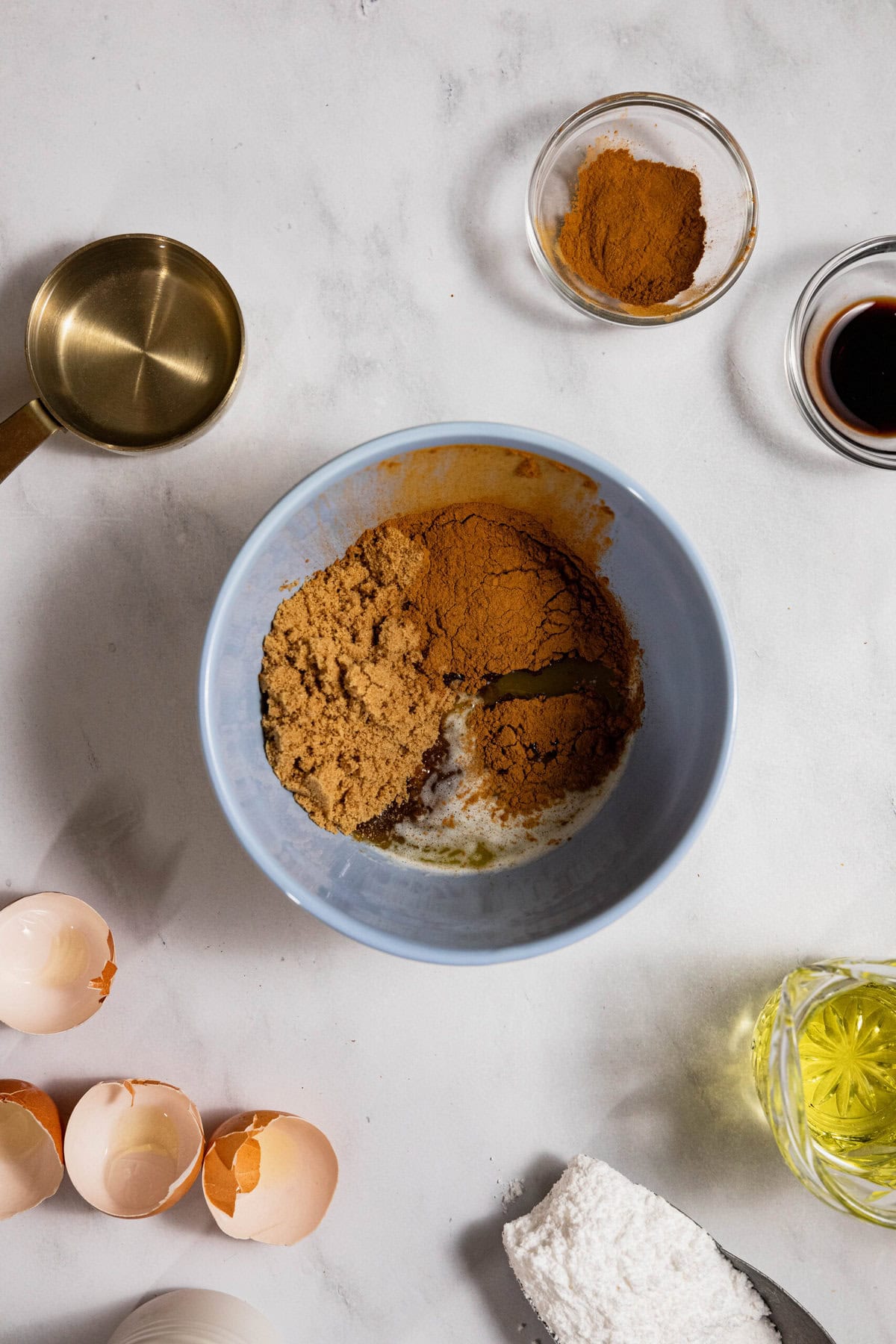 Baking ingredients on a counter: a bowl with brown sugar and cinnamon, a cracked egg, flour, vanilla extract, and a measuring cup.