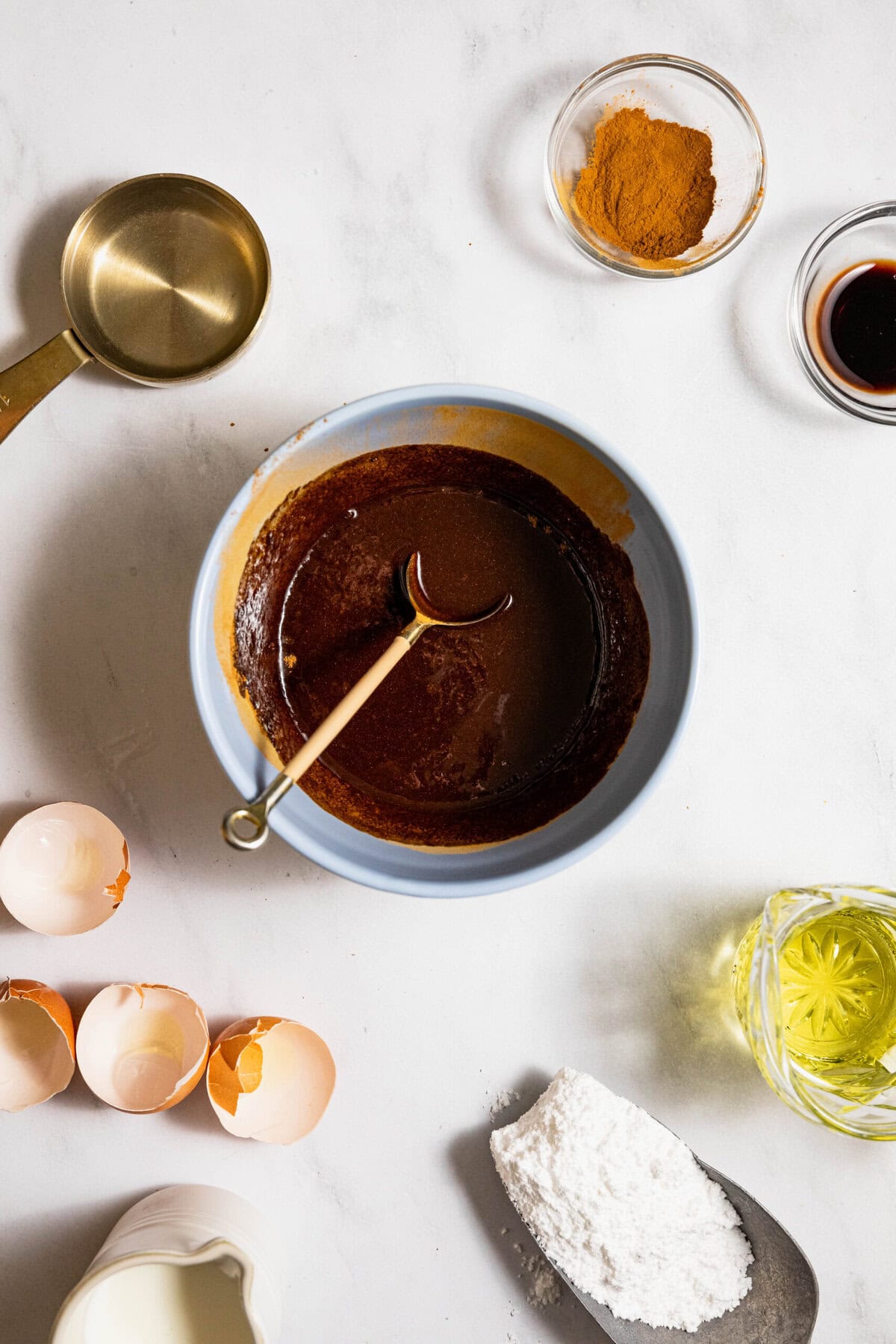 Ingredients for baking with a bowl of mixed batter in the center, surrounded by cracked eggshells, cinnamon, vanilla extract, measuring cup, oil, milk, and flour on a white surface.