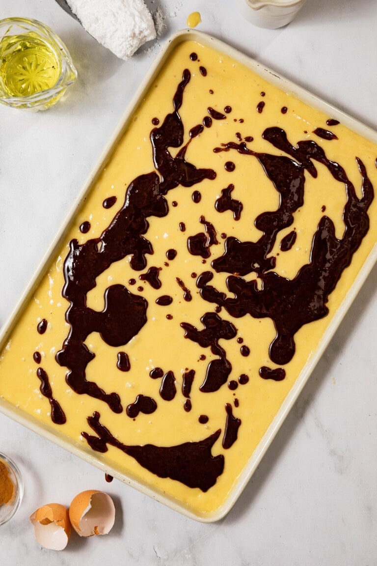 A baking tray filled with yellow batter topped with swirls of chocolate batter. Nearby are eggshells, a glass of oil, and a small flour-covered cloth.