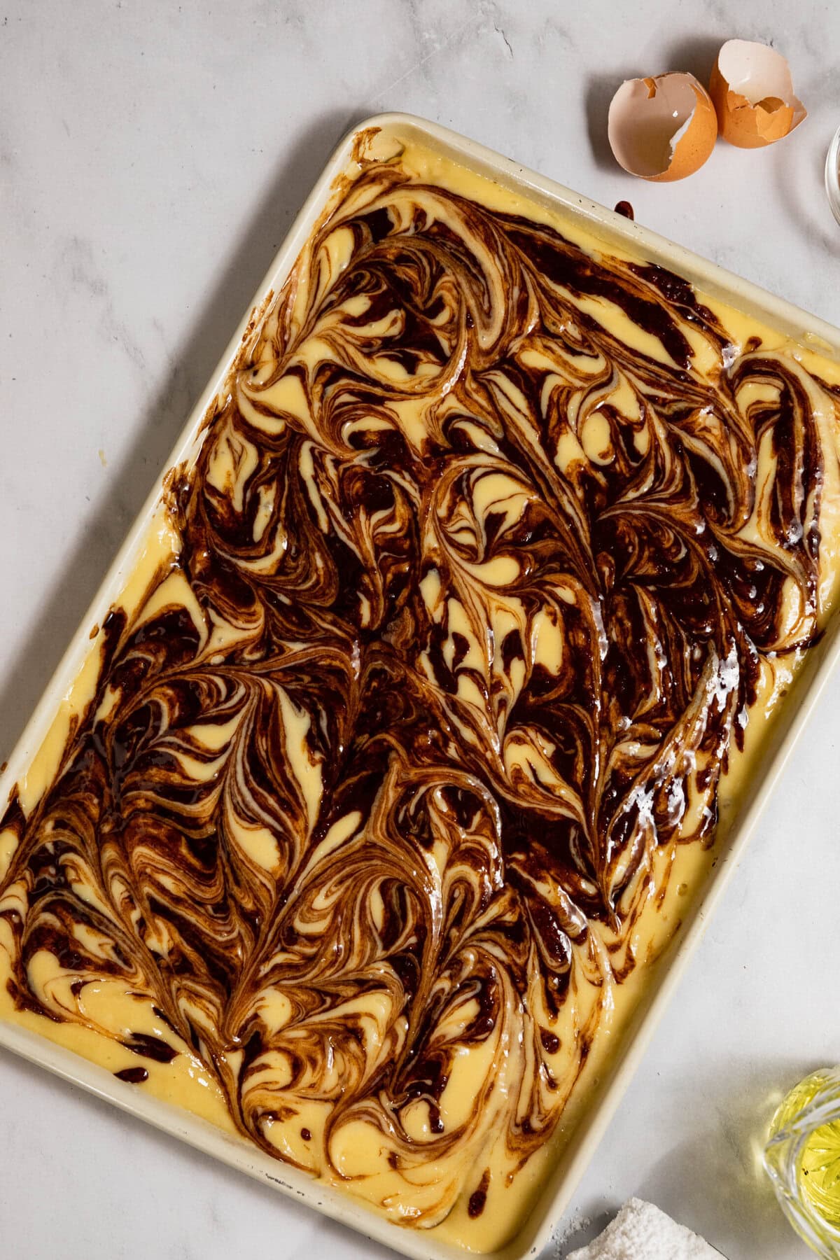 A baking tray filled with marbled chocolate and vanilla batter on a countertop, with egg shells in the background.