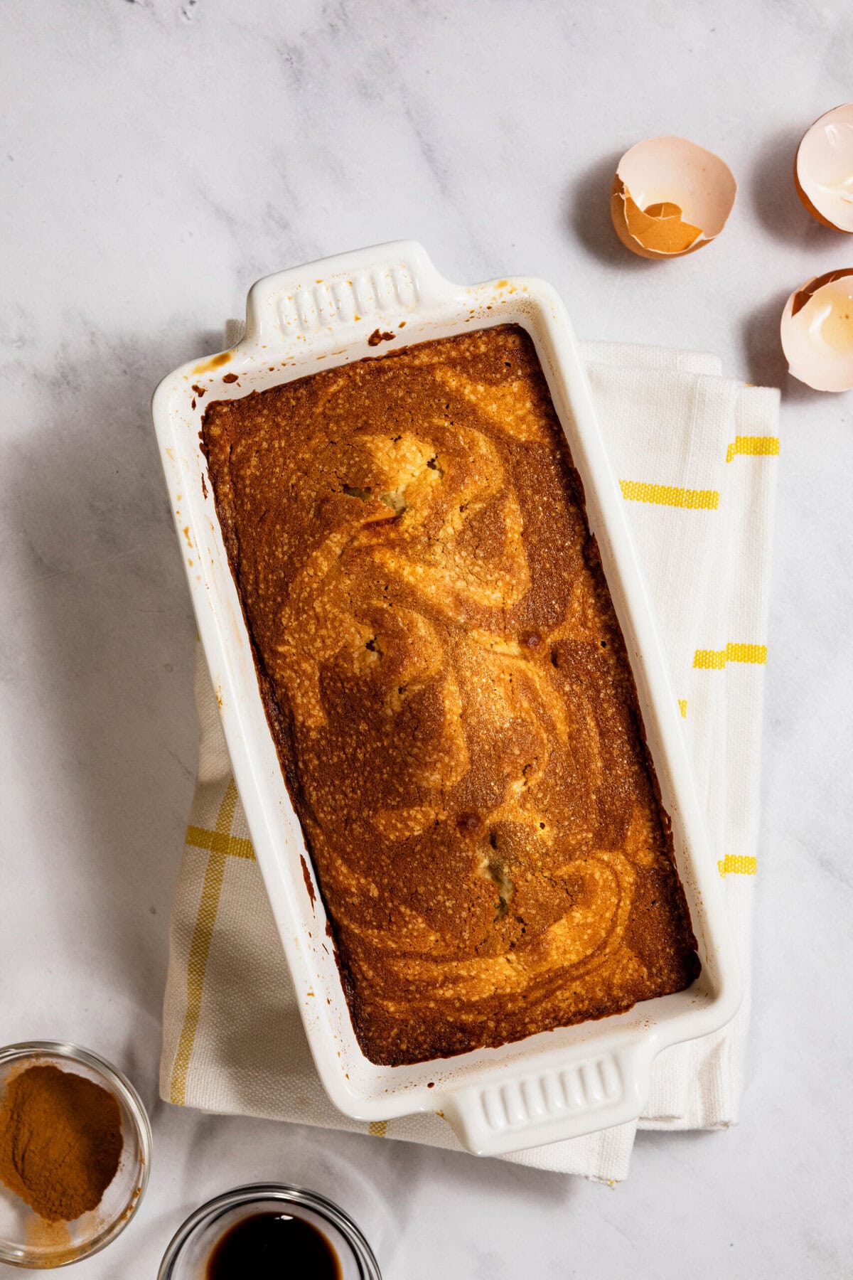 A rectangular loaf of baked bread in a white dish sits on a towel. Broken eggshells and small bowls of ingredients are nearby on a marble surface.