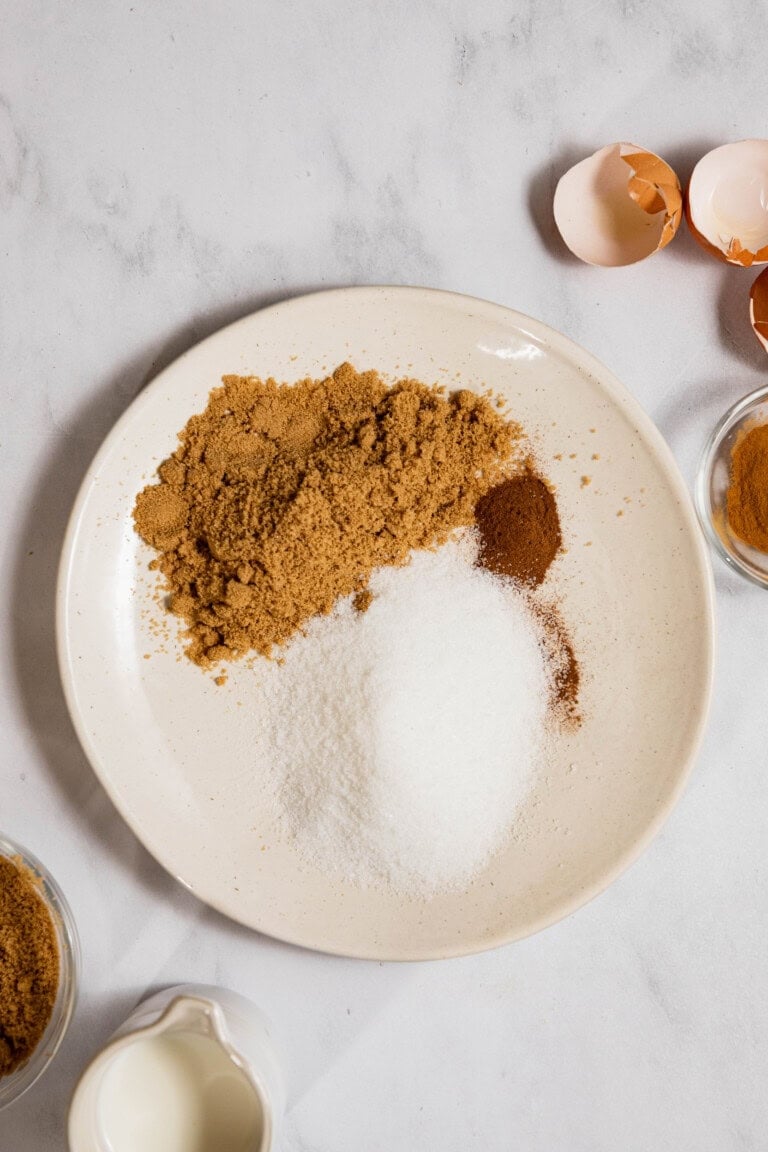 A plate with brown and white sugar, cinnamon, and ground spices on a marble surface. Eggshells, cinnamon, and milk are nearby.