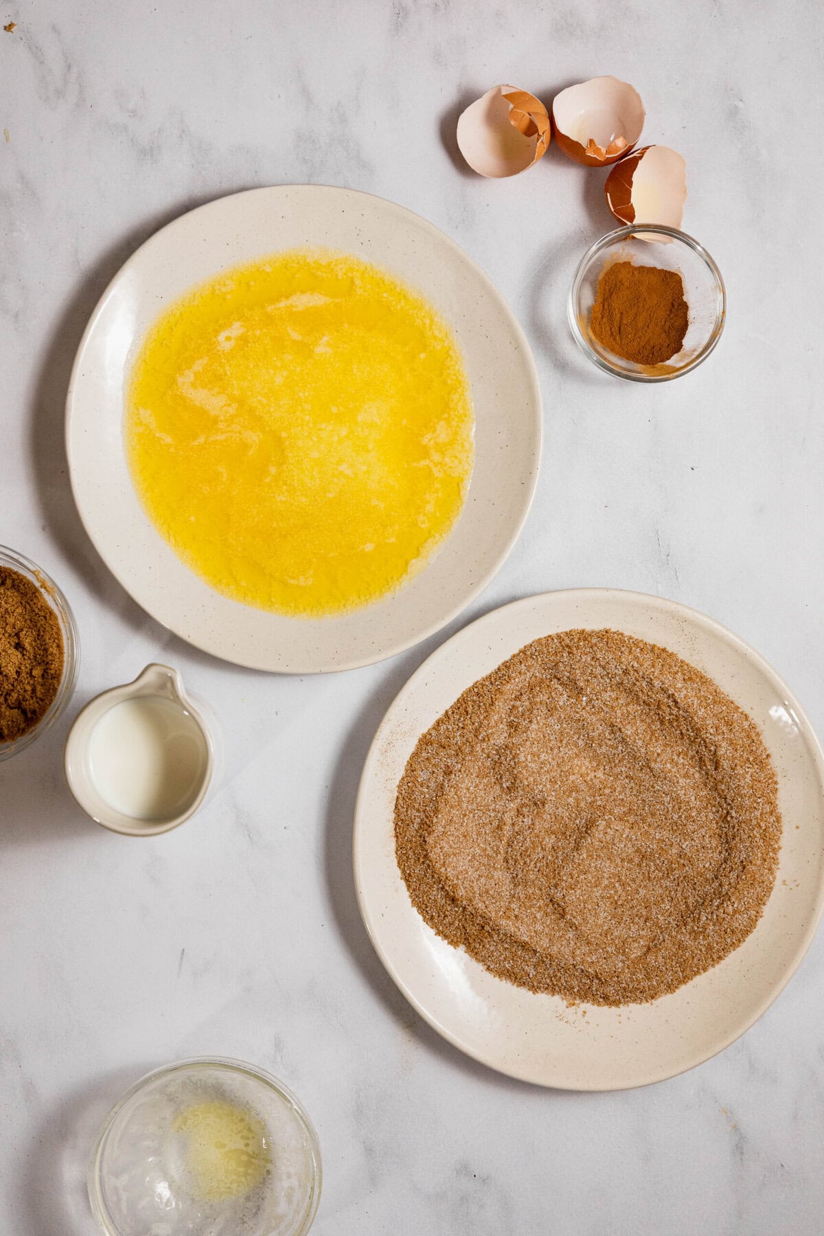 Ingredients laid out for baking: melted butter in a bowl, cinnamon and sugar mixture, brown sugar, milk, cracked eggshells, and a small bowl of cinnamon on a white surface.