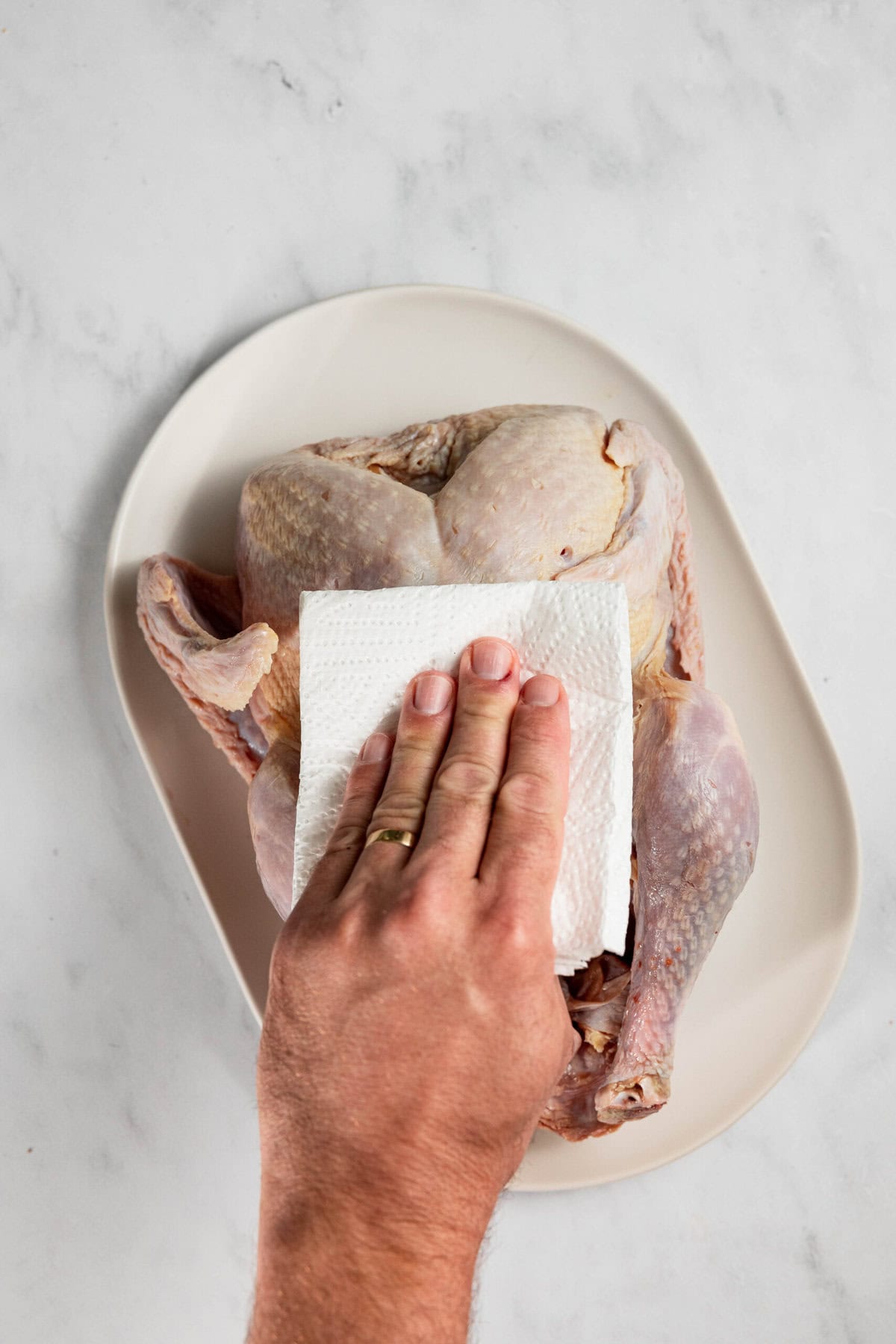 A hand using a paper towel to pat dry a raw chicken on a white plate.