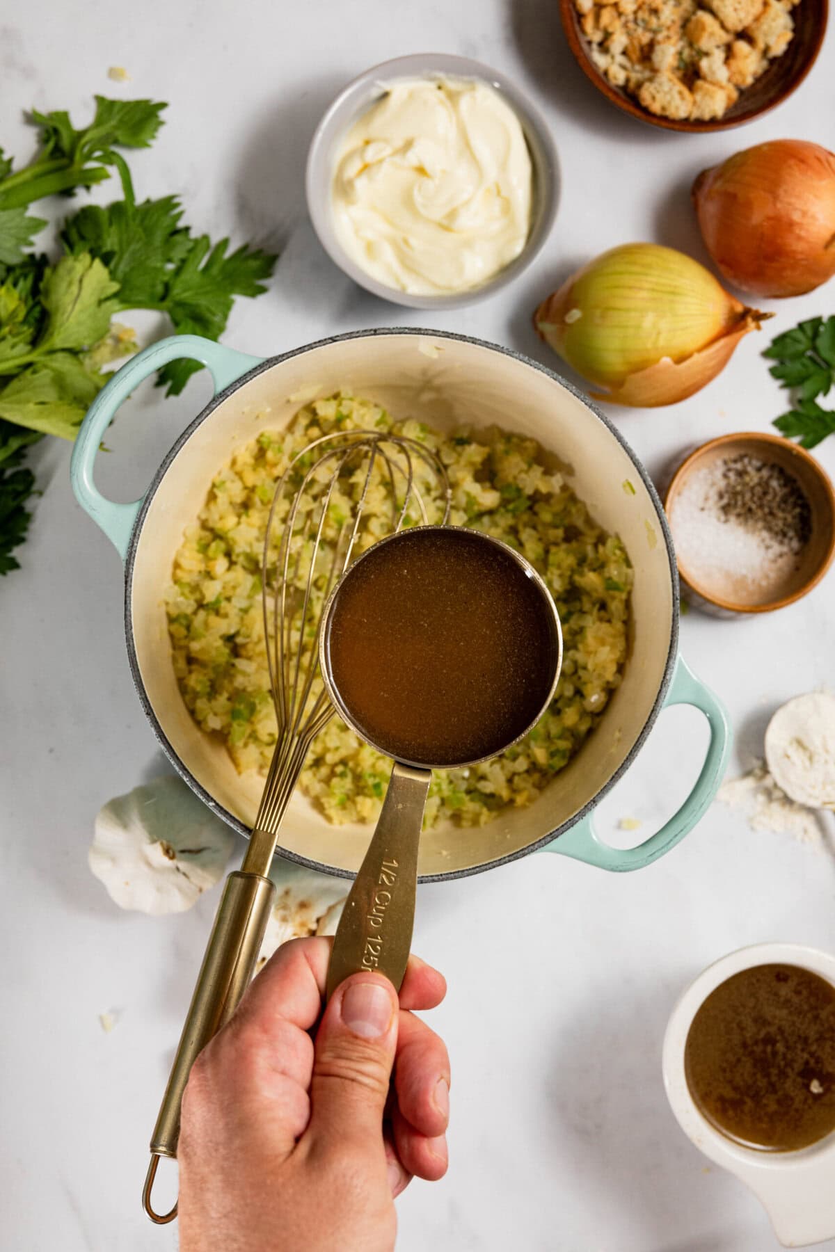 A hand holds a measuring cup with broth over a pot of chopped vegetables. Surrounding ingredients include mayonnaise, bread crumbs, onions, celery, salt, pepper, flour, and parsley on a marble surface.