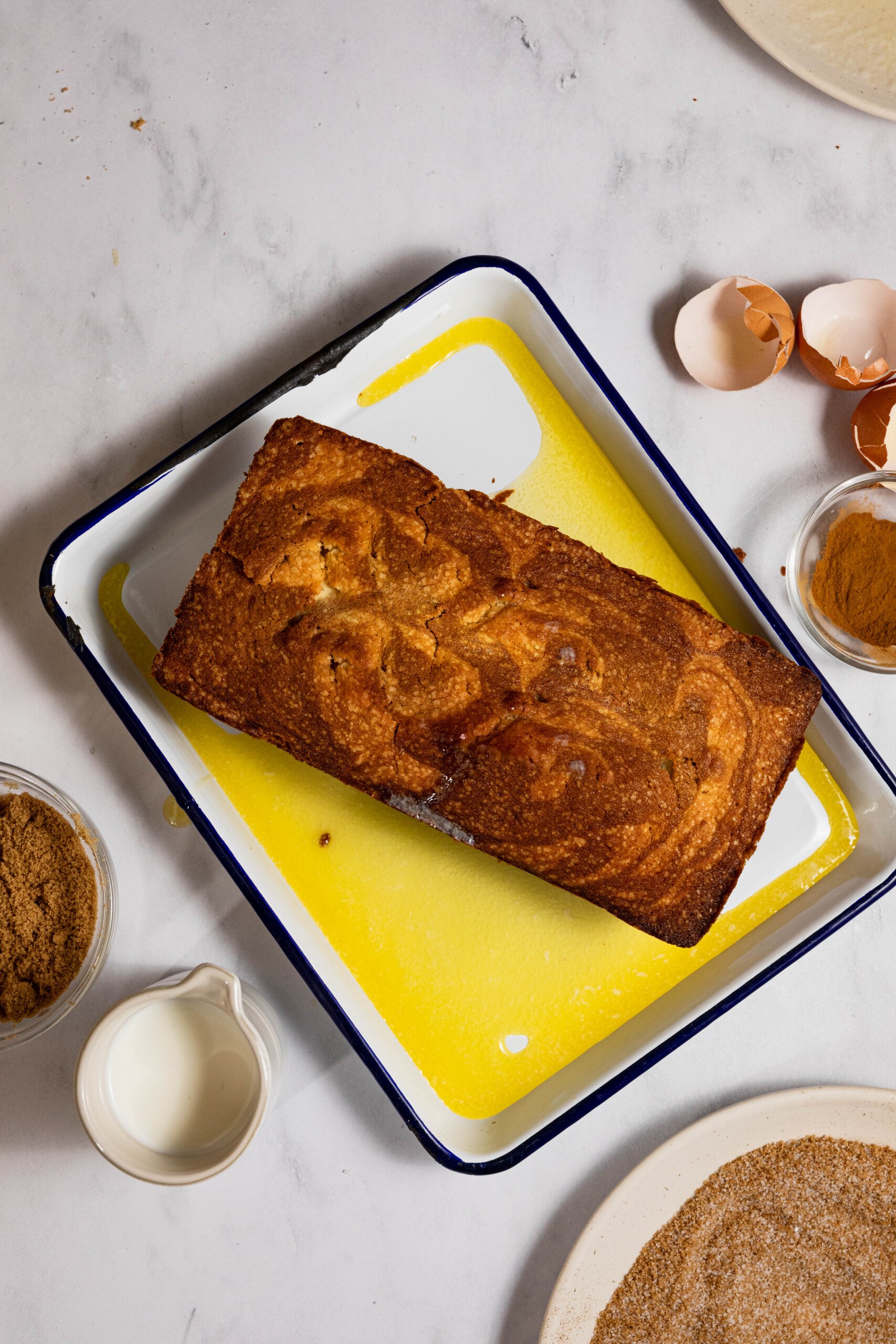 A loaf of golden-brown bread on a rectangular pan, surrounded by milk, cinnamon, brown sugar, eggshells, and a plate with sprinkled sugar on a marble countertop.