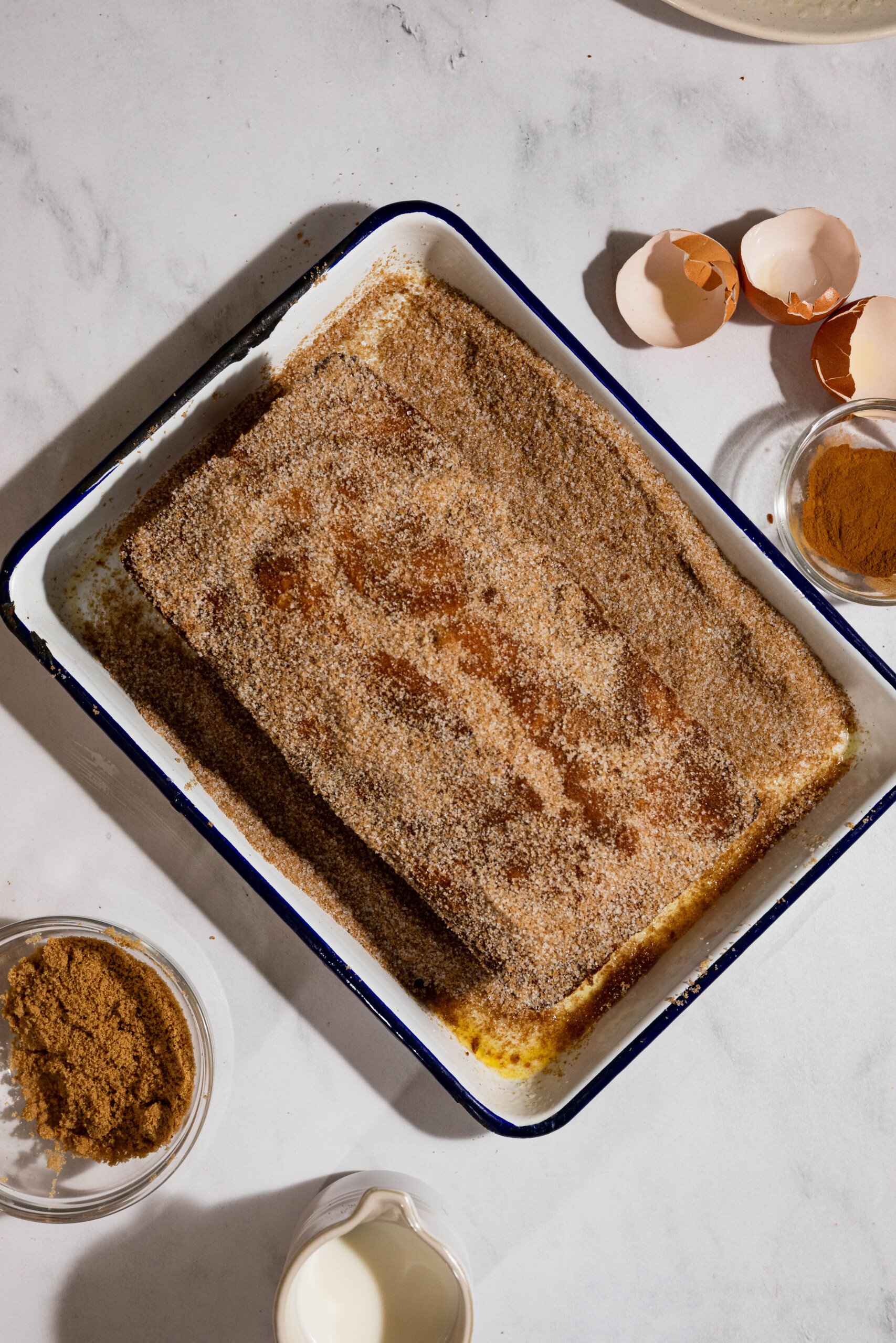 A baking tray with a rectangular dessert covered in a brown crumb mixture, surrounded by eggshells, brown sugar, cinnamon, and a cup of milk on a marble countertop.