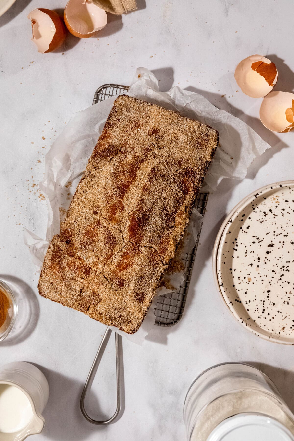 A loaf cake on parchment paper rests on a wire rack, surrounded by eggshells, plates, and small dishes.