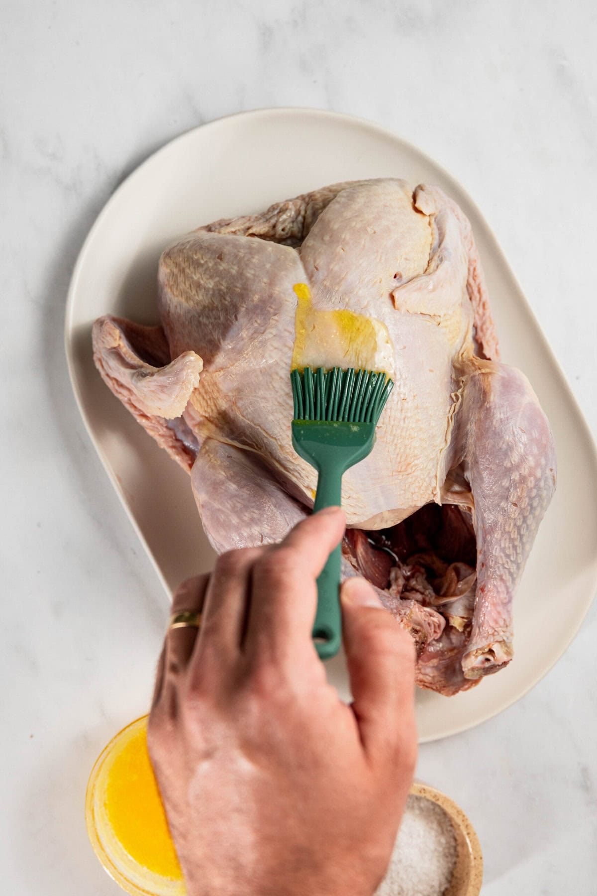 A raw whole chicken is placed on a white plate. A hand holds a green brush, applying melted butter to the chicken. A small bowl of salt is visible nearby.