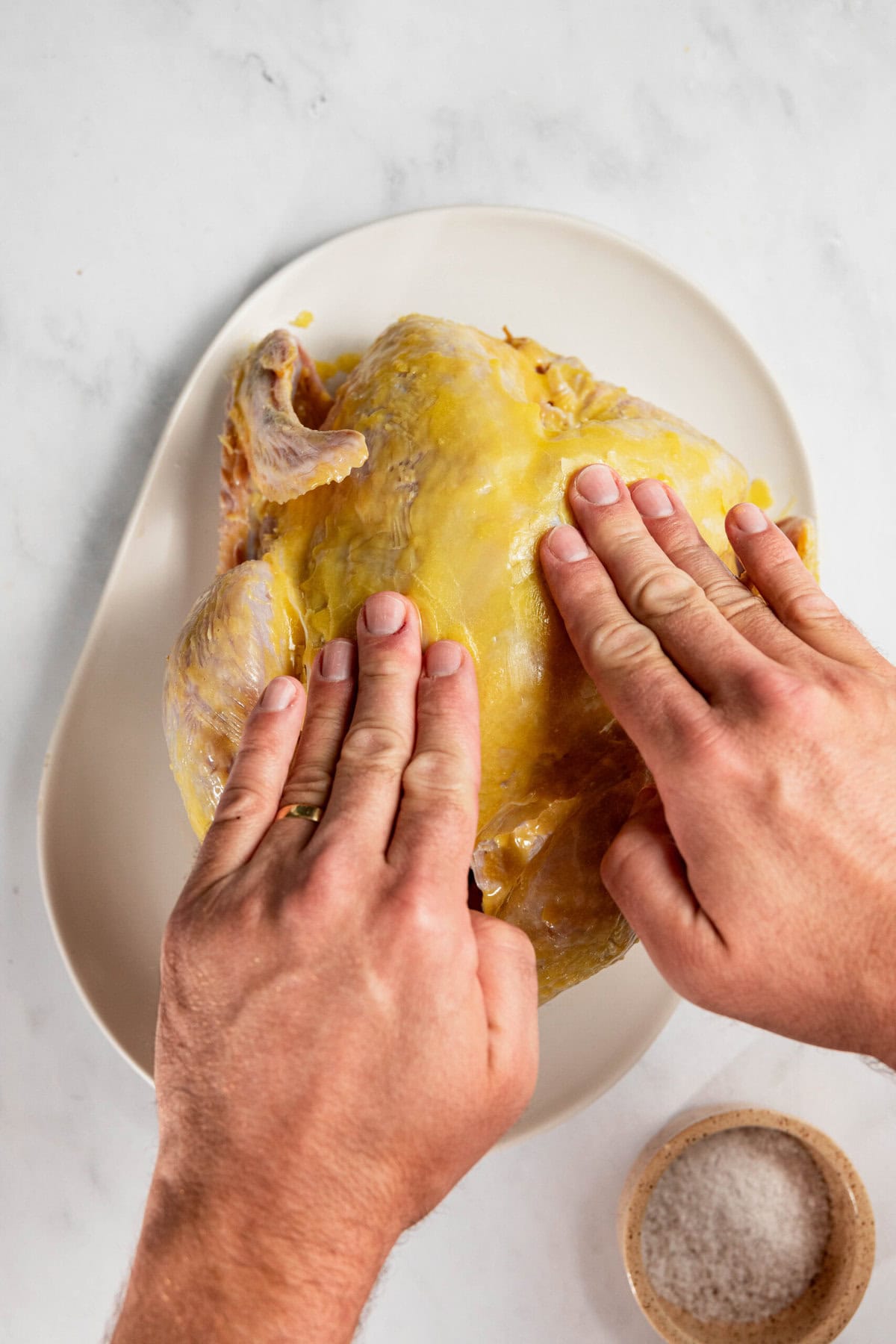 Hands applying seasoning to a raw chicken on a white plate with a small bowl of salt nearby.