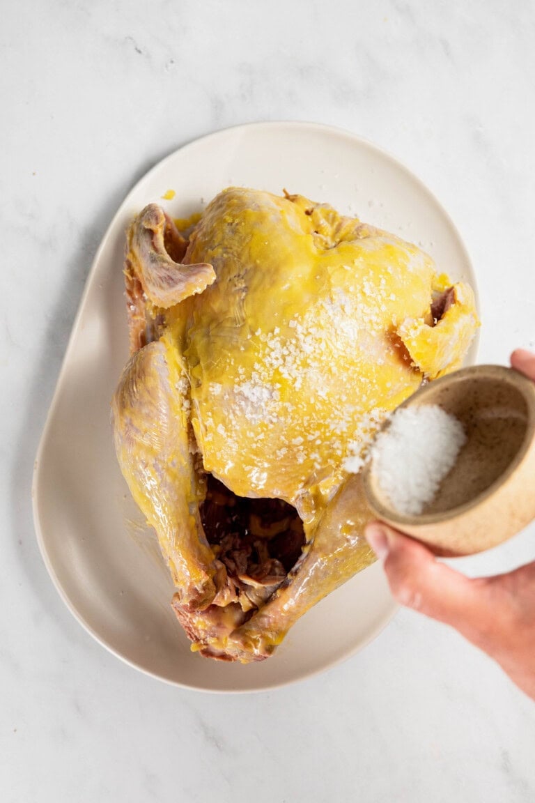 Raw whole chicken on a plate being seasoned with salt from a small bowl.