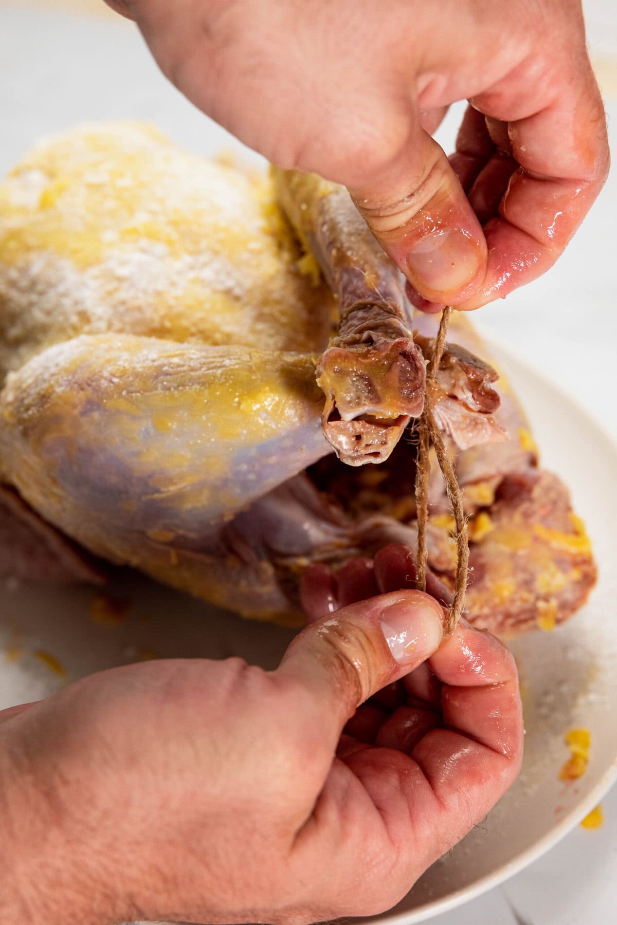 Hands tying the legs of a raw chicken with twine on a plate.