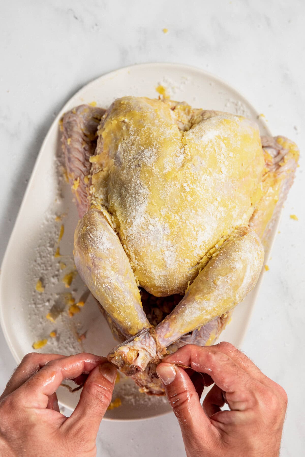 Hands preparing a raw whole chicken coated with seasoning on a white plate.