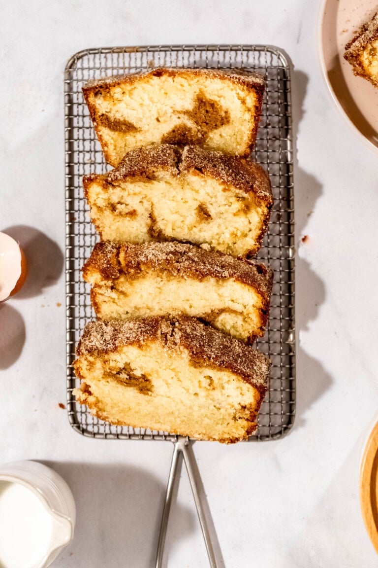 Sliced cinnamon swirl coffee cake on a wire cooling rack, surrounded by a small pitcher of milk and an empty eggshell.