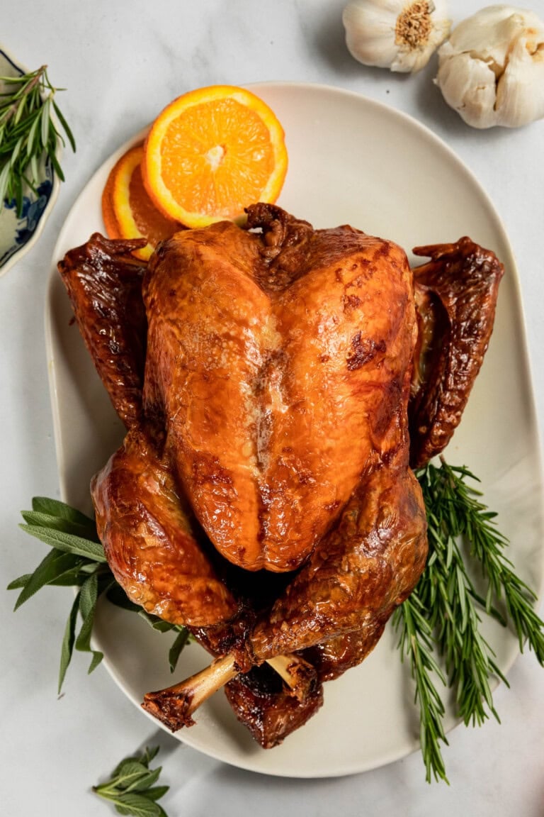 A roasted turkey on a white platter garnished with fresh rosemary, sage, and orange slices, with garlic bulbs nearby.