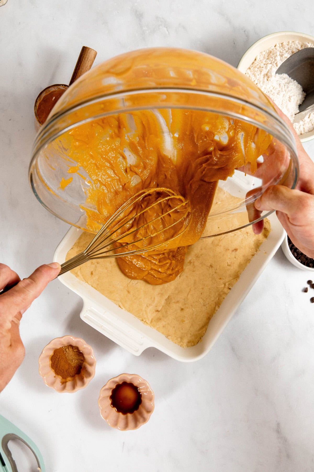 A person pours pumpkin batter from a glass bowl into a baking dish filled with dough, surrounded by small bowls of spices and flour on a white surface.