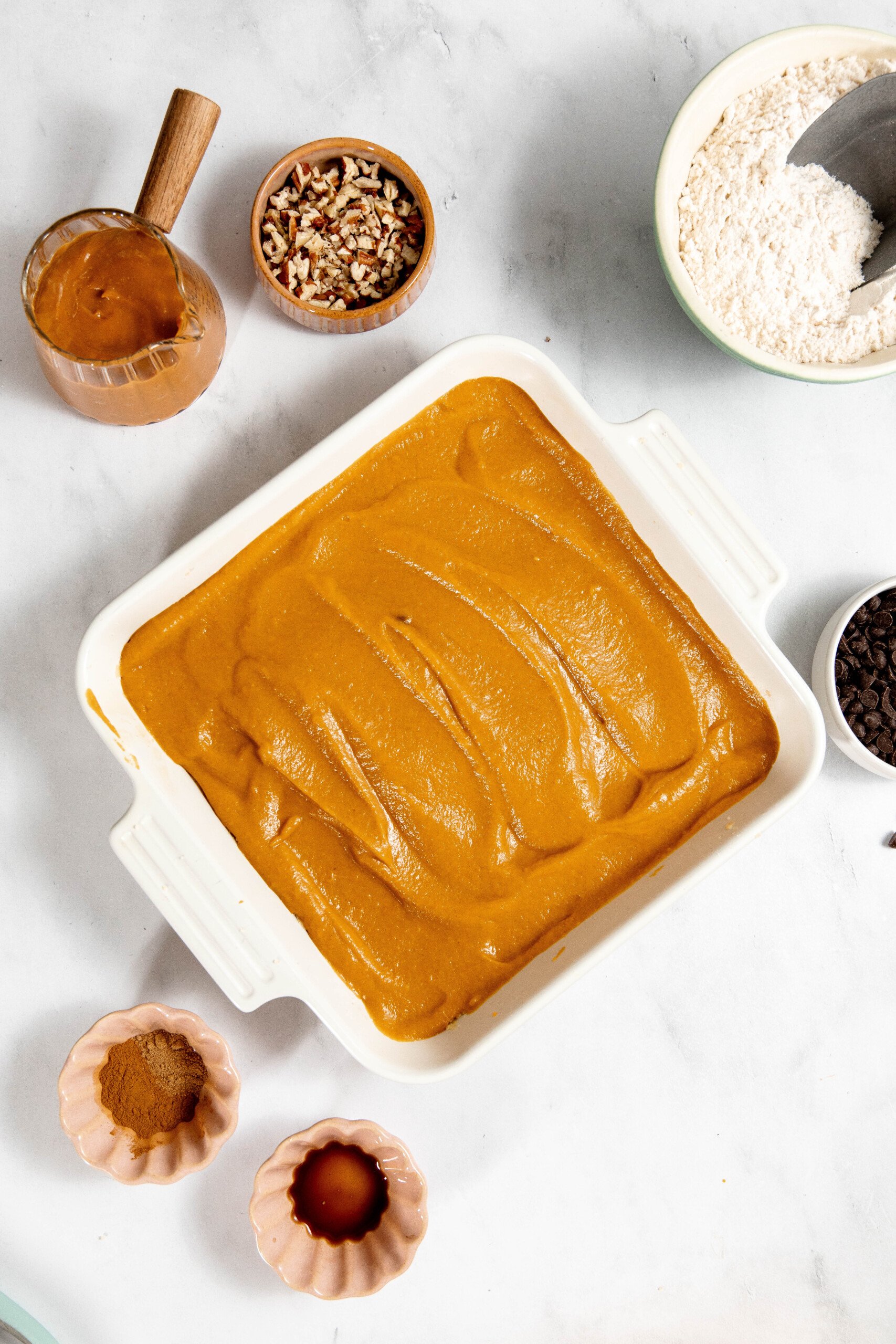 A baking dish filled with pumpkin batter is surrounded by ingredients like flour, cinnamon, vanilla extract, chopped nuts, chocolate chips, and a jar of wet ingredients on a marble surface.