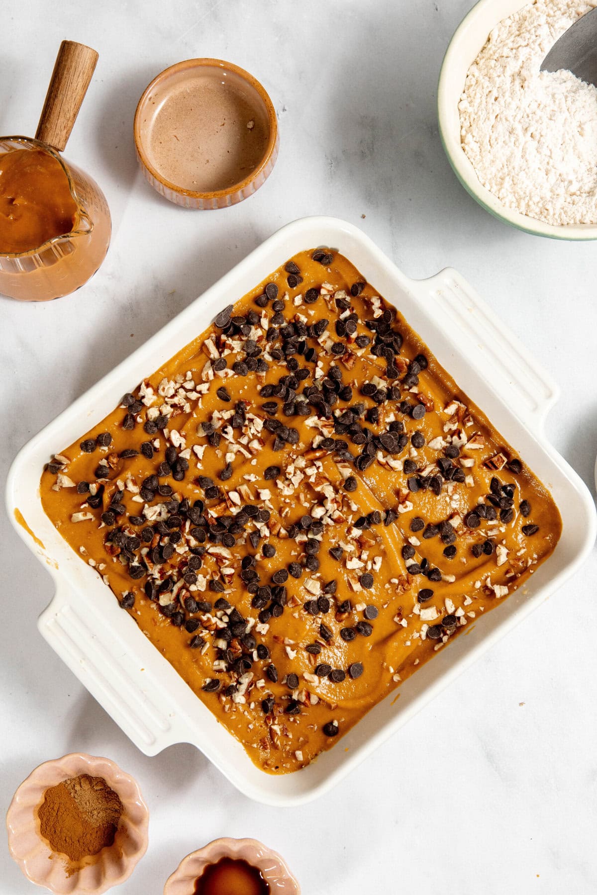 Baking dish with dough topped with chocolate chips and nuts. Surrounded by bowls of ingredients on a marble surface.