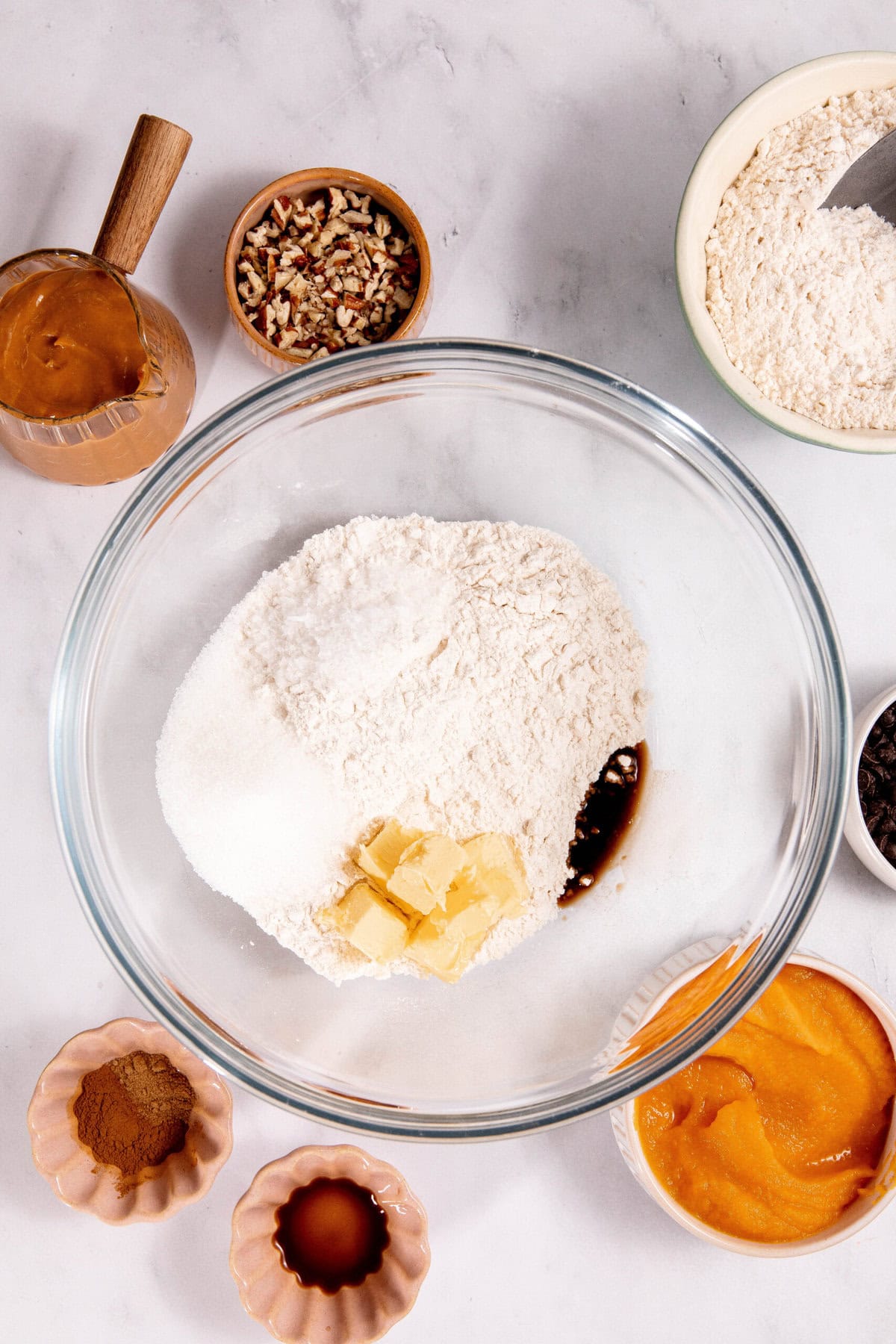 Baking ingredients arranged on a countertop, including flour, sugar, butter, vanilla, pumpkin puree, spices, nuts, and chocolate chips in various bowls and containers.