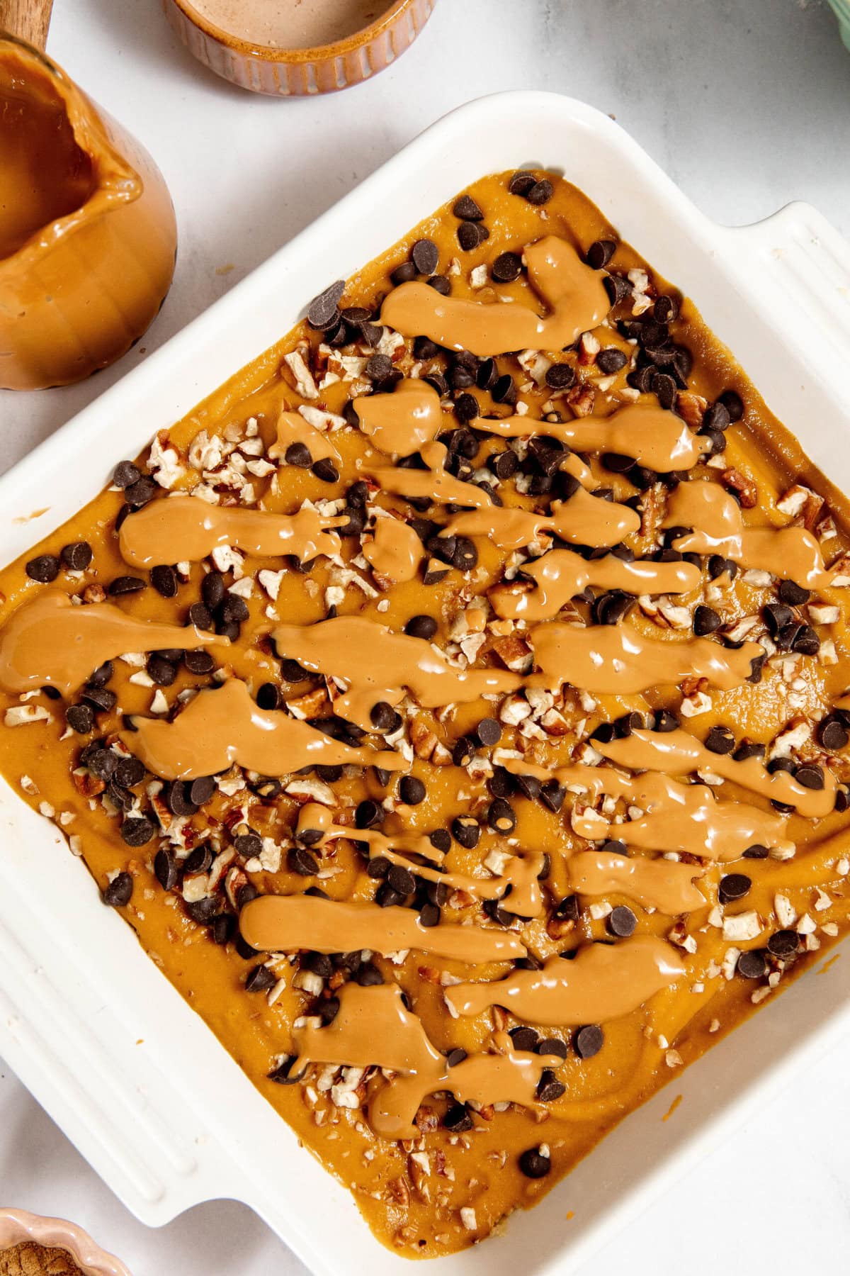 A square pan filled with a pumpkin dessert topped with chocolate chips, chopped nuts, and drizzled caramel.