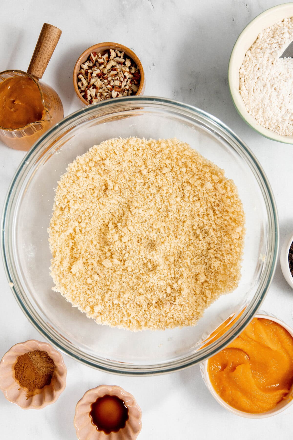 A large glass bowl with crumbly dry ingredients, surrounded by smaller bowls containing pecans, flour, spices, vanilla extract, pumpkin puree, and caramel.