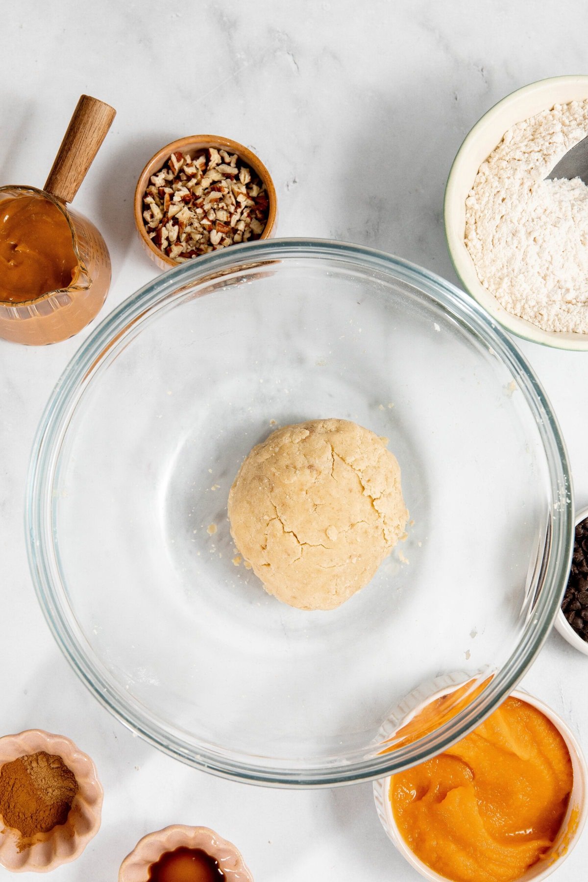 Bowl with dough surrounded by flour, spices, nuts, pumpkin puree, caramel, and chocolate chips on a marble surface.