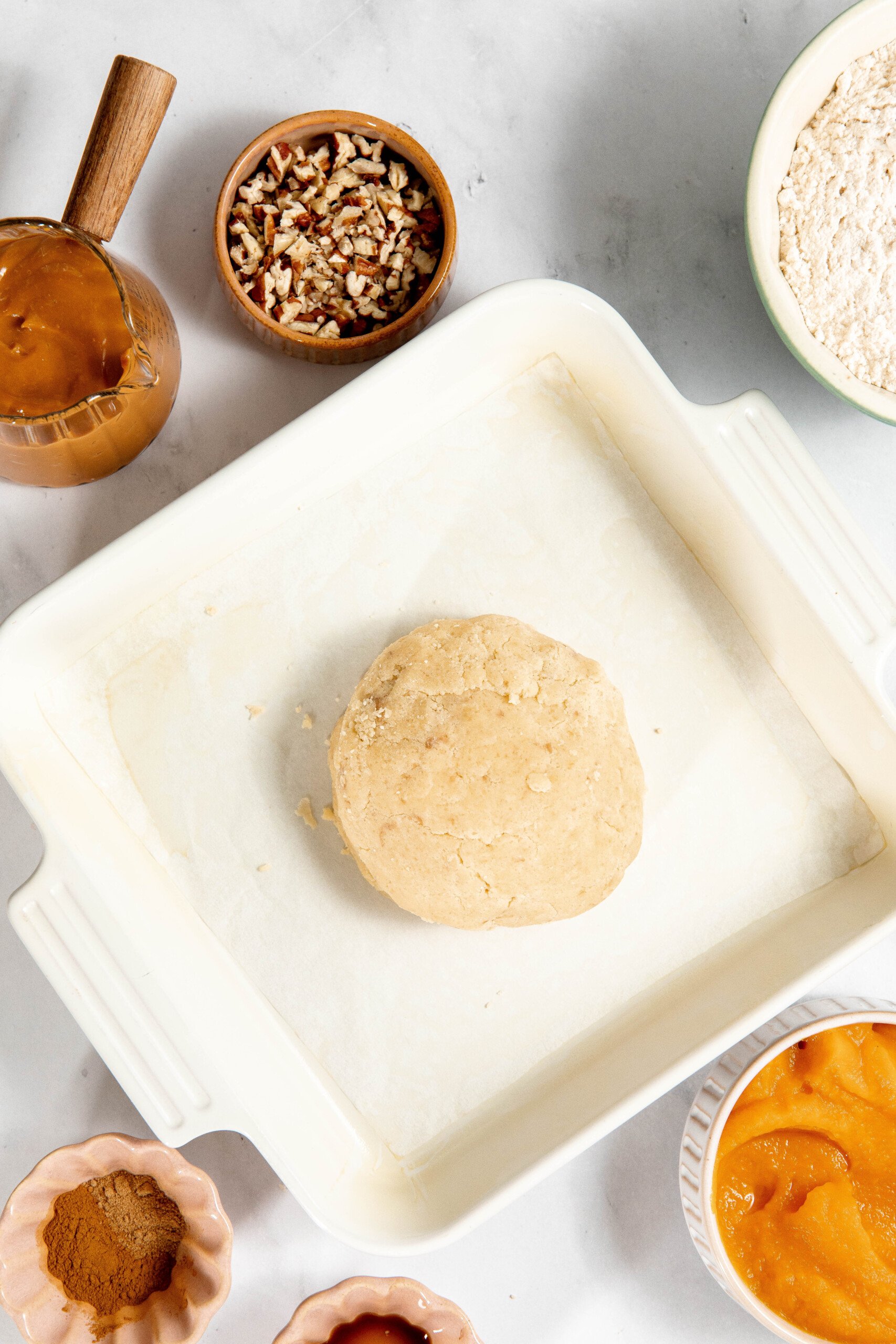 Baking setup: dough in a square dish, surrounded by bowls of nuts, flour, pumpkin puree, cinnamon, and brown sugar on a marble surface.