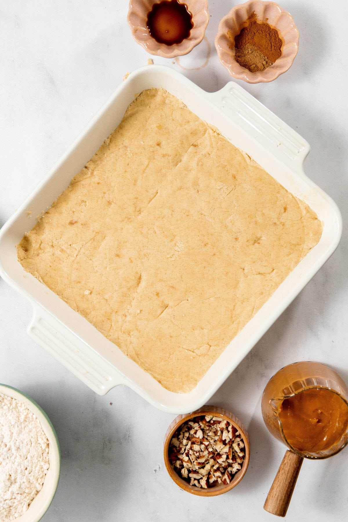 Square baking dish with dough, surrounded by bowls of flour, vanilla, cinnamon, chopped pecans, and caramel on a marble surface.