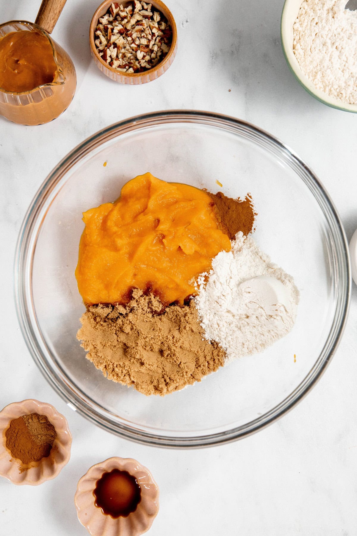 Bowl with pumpkin puree, brown sugar, flour, and spices. Nearby are bowls of vanilla extract, pecans, and cinnamon.