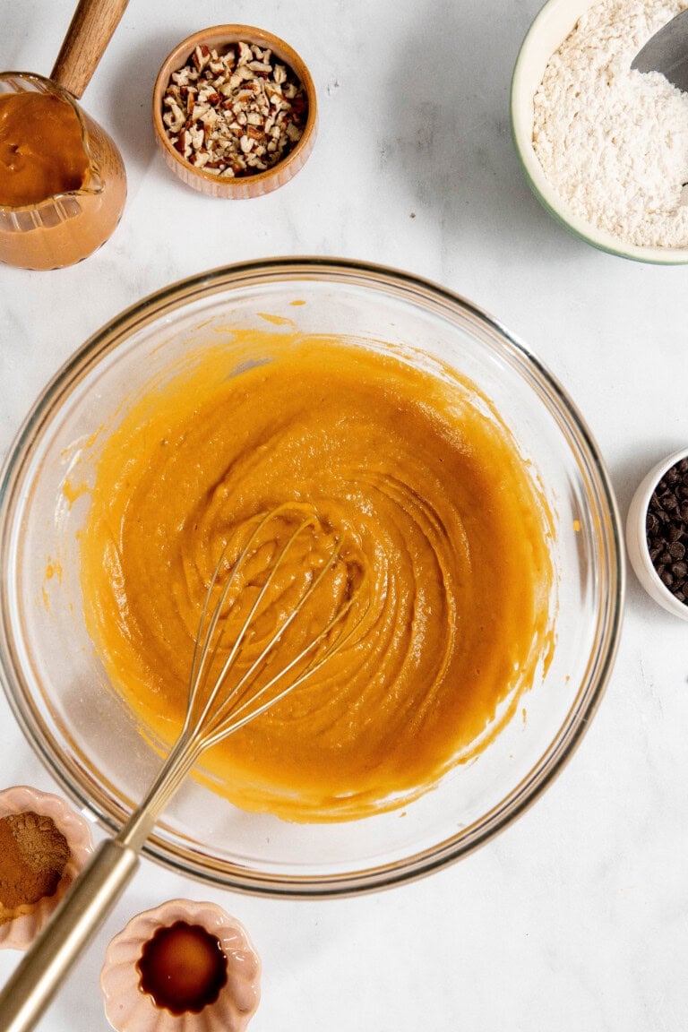 A glass bowl with pumpkin batter and a whisk. Nearby are bowls of flour, pecans, chocolate chips, vanilla, and caramel on a white surface.