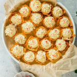 A plate of cream-topped tarts with red and white sprinkles, placed on parchment paper. A small dish with peppermint candies is nearby, on a light-colored surface.